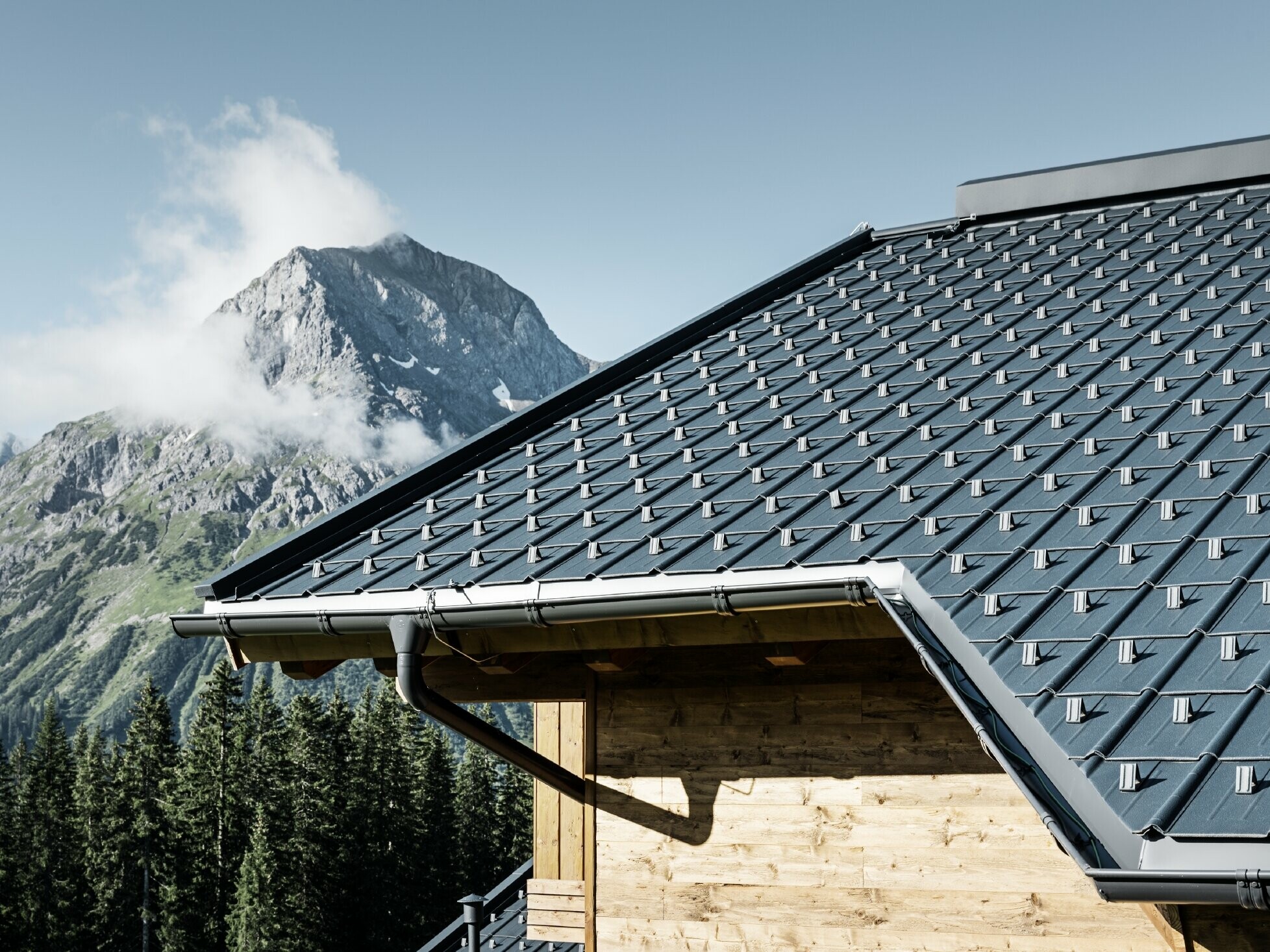 PREFA-tagplade i alpin anvendelse – tagplade i antracit opsat på hytte i Lech am Arlberg med bjerge i baggrunden; PREFA-tagrende i antracit; træfacade