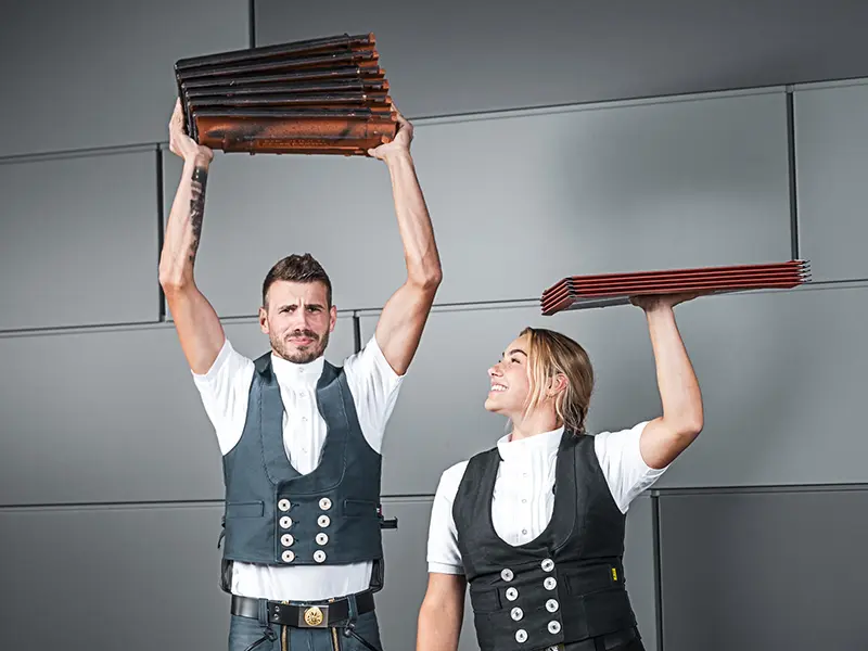 På billedet ses to personer, der hver holder en stak tagdækningsmateriale i hånden. Personer til venstre holder en stak tagsten over hovedet, mens personen til højre holder en stak aluminiumsplader over hovedet. Begge er klædt i traditionelt håndværkerudstyr med vest og hvid skjorte. Baggrunden er moderne og minimalistisk med grå vægge. Begge personer virker koncentrerede og smiler.