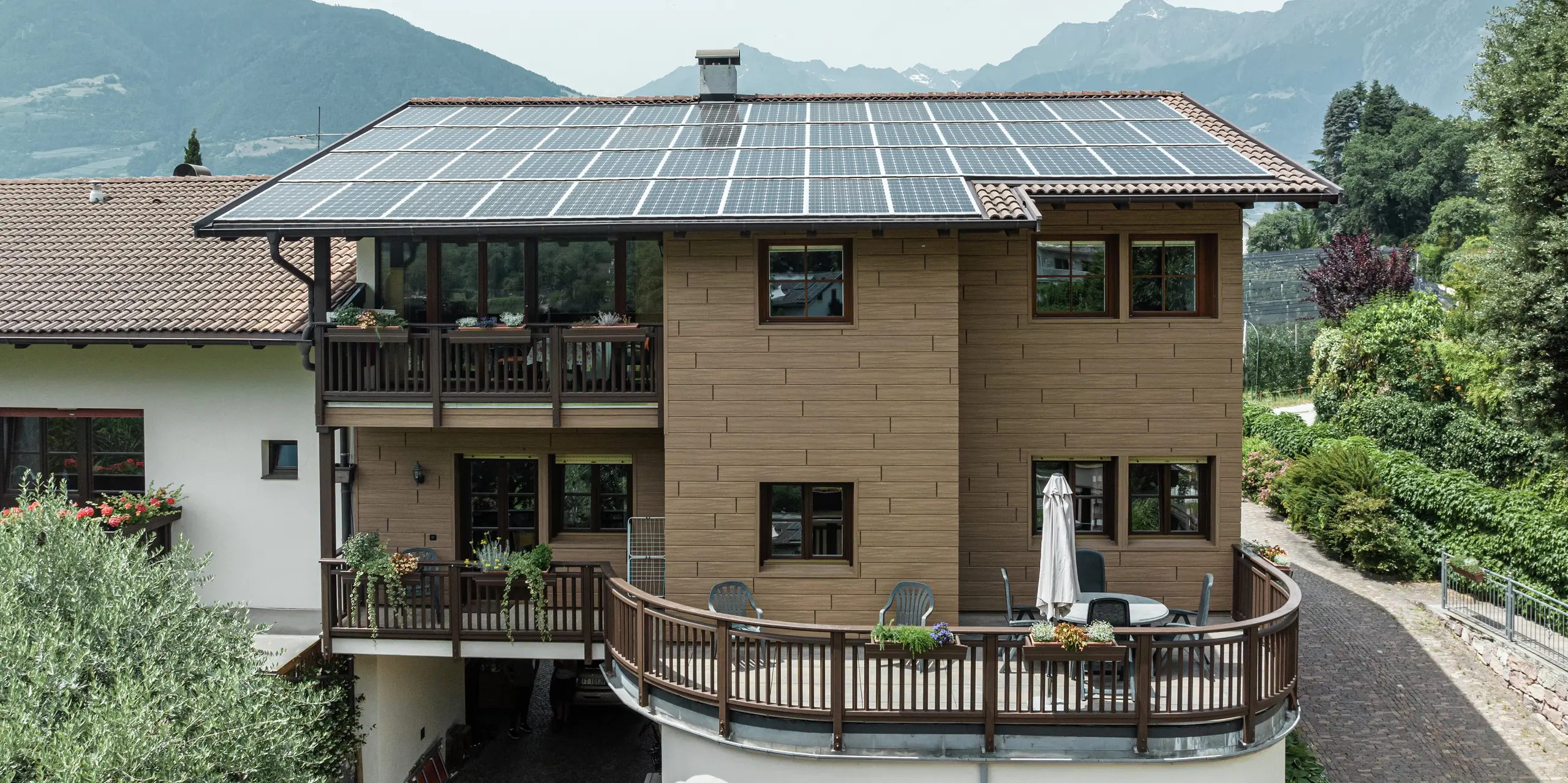 Sanierte Ferienwohnungen Prechtlhof in Meran, Südtirol, mit PREFA Sidings in Walnussbraun und einer großflächigen Photovoltaikanlage am Dach. Die wartungsarme Aluminiumfassade in Holzoptik verbindet Nachhaltigkeit mit traditioneller Ästhetik und fügt sich perfekt in die alpine Landschaft ein.