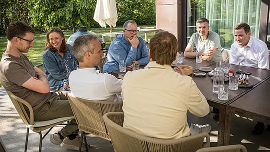 PREFARENZEN 2027 Expertendialog im sonnendurchfluteten Außenbereich einer Winzerresidenz: Architekten und Designexperten sitzen um einen großen Tisch, tief in Diskussionen vertieft, während Glaswaren und kleine Häppchen auf dem Tisch arrangiert sind. Die Szene zeigt lebendige Interaktion und konzentrierte Aufmerksamkeit, flankiert von natürlichem Grün und Schatten der Bäume. PREFA Aluminiumprodukte im Hintergrund stehen für langlebige, ästhetisch ansprechende Dächer und Fassaden. Das Bild vermittelt, wie PREFA Design, Funktion und Nachhaltigkeit kombiniert und kreative Diskussionen zwischen Architekten inspiriert.