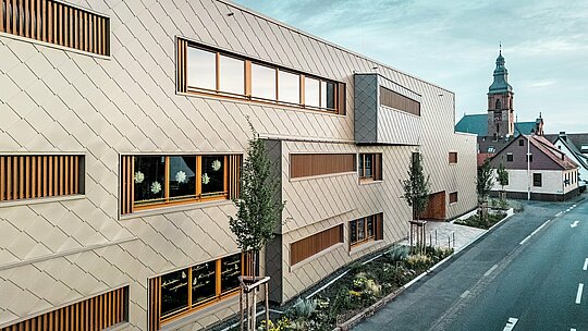 Die Seitenansicht der Kindertagesstätte in Alzenau zeigt das moderne Hybridgebäude im spannungsvollen Kontrast zur historischen Stadtstruktur mit der spätgotischen Burgkirche im Hintergrund. Die Fassade aus bronzefarbenen PREFA Wandrauten 44 × 44 erzeugt ein elegantes, rhythmisches Muster und hebt sich selbstbewusst vom Umfeld ab – ohne sich aufzudrängen. Großzügige Fensteröffnungen mit vertikalen Holzlamellen und die strukturierte Begrünung entlang des Gebäudes unterstreichen das nachhaltige Gestaltungskonzept. Die Kombination aus innovativer Aluminiumfassade, ökologischer Bauweise und städtebaulichem Feingefühl macht das Projekt zu einem Paradebeispiel für verantwortungsvolle Architektur zwischen Tradition und Zukunft.