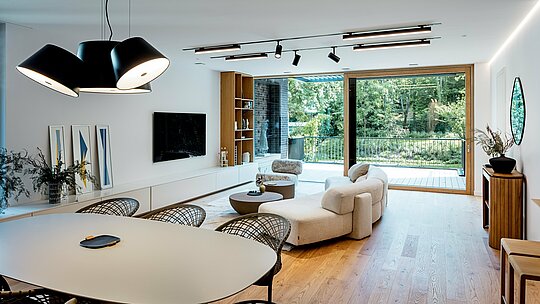 Die Aufnahme zeigt den lichtdurchfluteten Wohn‑ und Essbereich der modernen Mehrgenerationen‑Villa in Budapest. Große Glasfronten öffnen den Raum zur begrünten Terrasse, während helle Holzböden, elegante Designmöbel und ein harmonisch abgestimmtes Beleuchtungskonzept eine warme Atmosphäre schaffen. Der offene Grundriss verbindet Küche, Essplatz und Wohnzimmer nahtlos miteinander. Die hochwertige Innenarchitektur ergänzt die klare Formensprache des Gebäudes, dessen äußere Struktur durch langlebige PREFALZ Aluminiumprodukte in P.10 Anthrazit geprägt wird. Der Raum vermittelt Großzügigkeit, Modernität und Wohnkomfort auf höchstem Niveau.