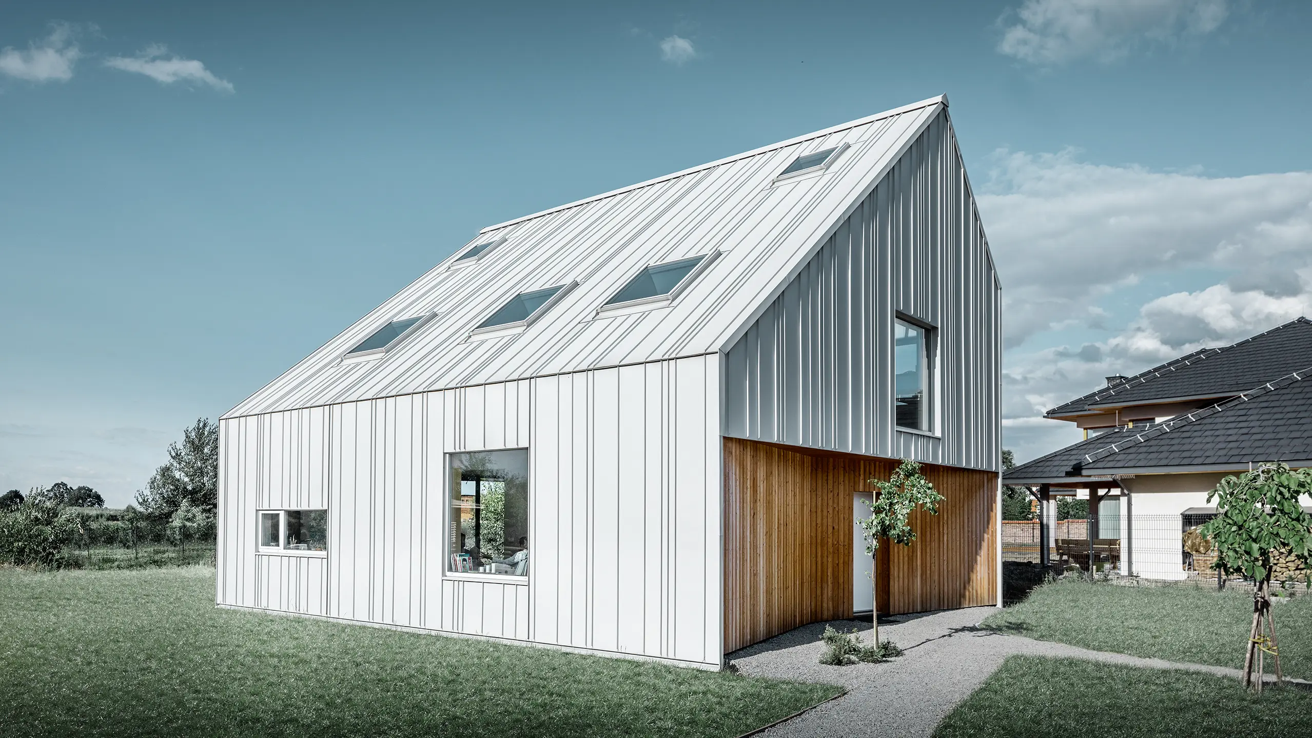 Moderne udformet enfamilieshus med sadeltag, hvor alle udvendige flader er beklædt med PREFALZ i Prefa-hvid. Derved strækker panelerne med forskellige bredder sig fra taget og videre over facaden. Der er sat flere tagvinduer i taget.
