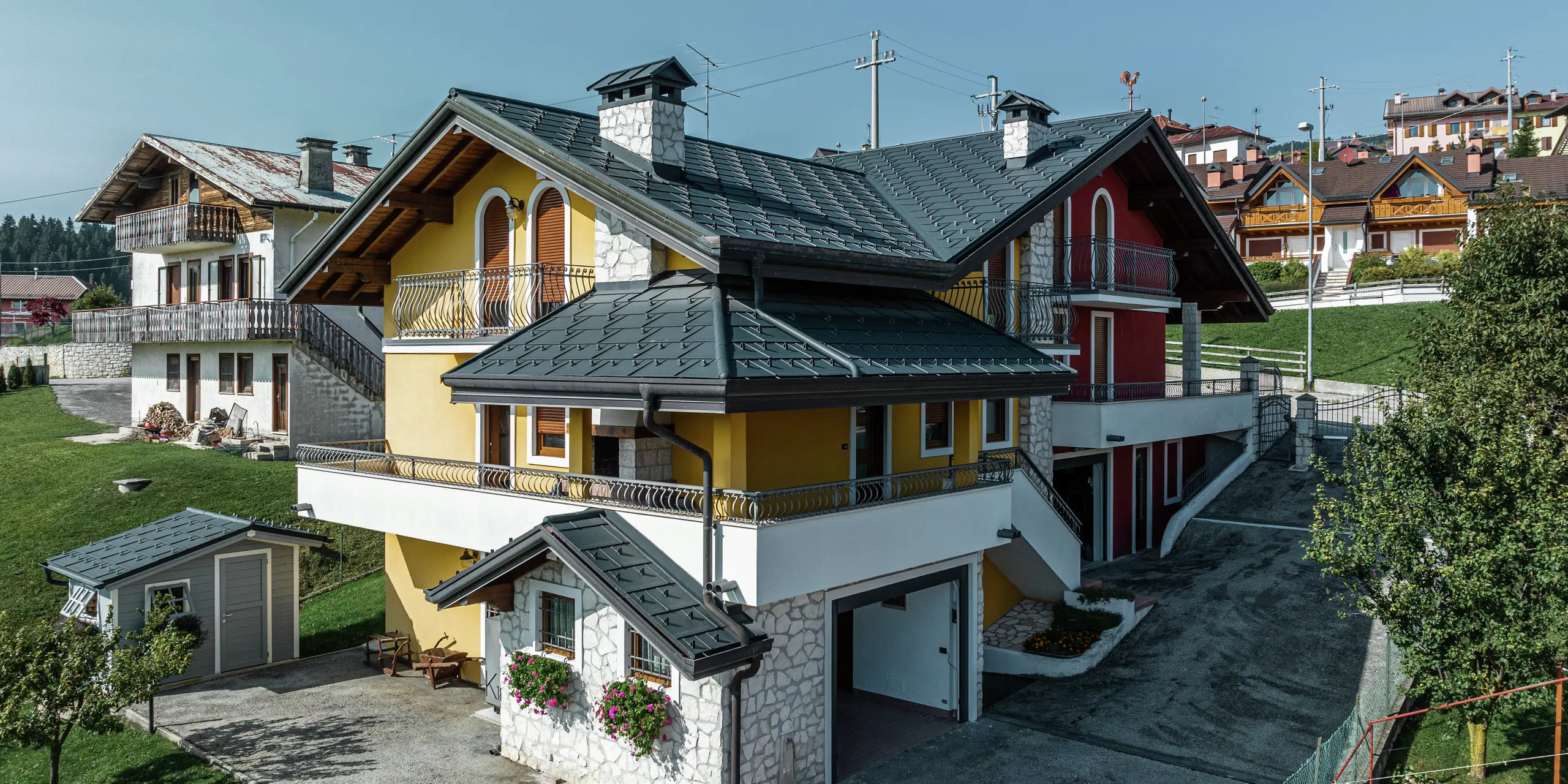 Einfamilienhaus in Gallio im alpinen Architekturstil mit modernem PREFA Blechdach in P.10 Dunkelgrau. PREFA Dachplatten R.16 sorgen für eine langlebige, witterungsbeständige und optisch ansprechende Dachlösung. Die harmonische Kombination aus gelb-roter Fassade, Natursteinelementen und Holzakzenten verleiht dem Haus einen einladenden Charakter. Die PREFA Dachentwässerung in P.10 dunkelgrau sorgt für eine zuverlässige Wasserableitung. Ein Paradebeispiel für ästhetische und funktionale Architektur in alpiner Umgebung.