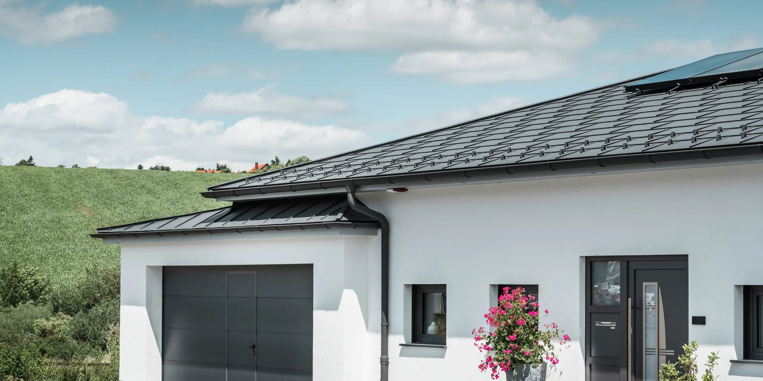 Billedet viser forsiden af et enfamiliehus i Ilmenau, Thüringen, med et elegant PREFA tag i P.10 mørk grå. Tagpladen R.16 giver huset et moderne udseende, mens det integrerede PREFA tagafvandingssystem med sin mørkegrå halvrunde tagrende og nedløbsrør sikrer effektiv afledning af regnvand. Garagen, der er udstyret med mørkegrå PREFALZ, passer harmonisk ind i den samlede æstetik og understreger kvaliteten og stilen af PREFA aluminiumsprodukter.