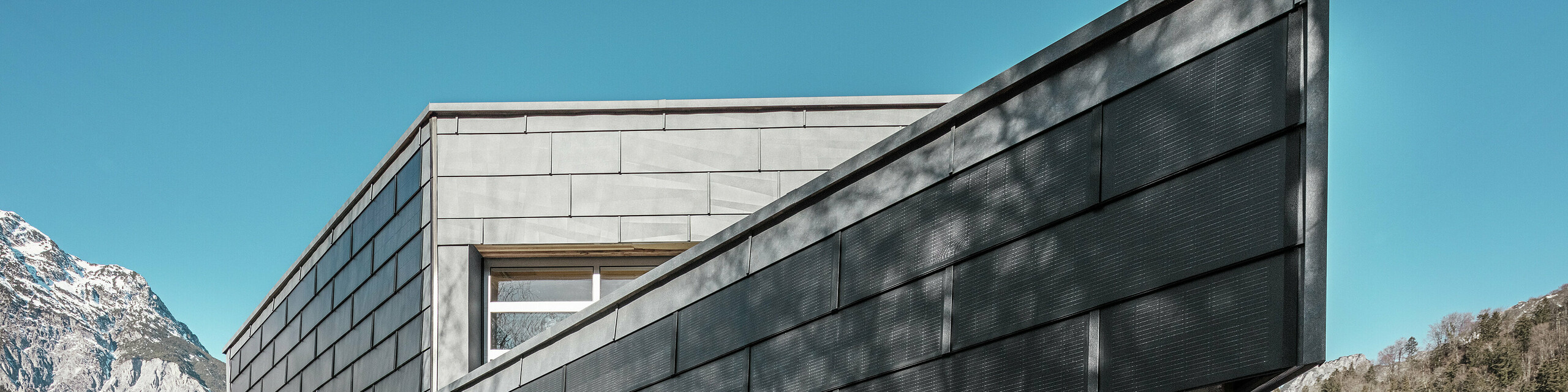 Das Bürogebäude der RÜMA Blechwerkstatt in Vorarlberg besticht durch seine moderne Fassade aus PREFA Solardachpaneelen und Sichtbeton. Umgeben von Bergen und Bäumen, deren Schatten sich an der Wand abzeichnen, erhebt es sich unter strahlend blauem Himmel. Die markante Gebäudekante und die PREFA Fassadenpaneele FX.12 unterstreichen den eindrucksvollen Eindruck zusätzlich.