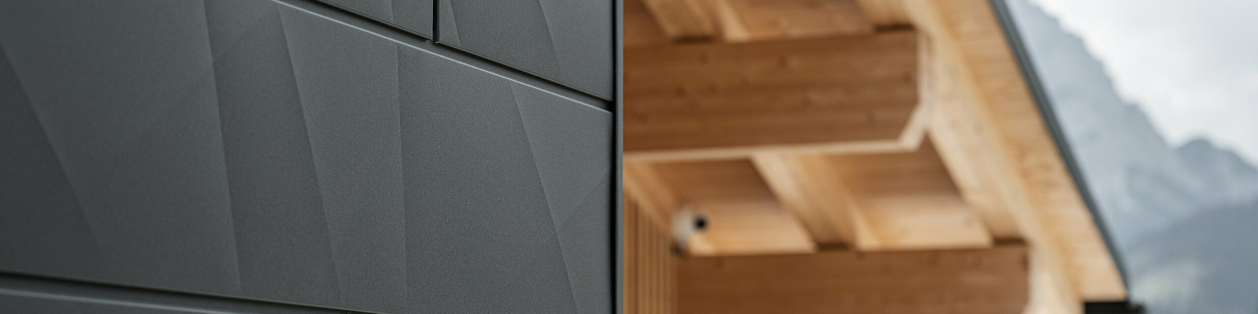 Nærbillede af Siding.X facadeplader i P.10 sort fra PREFA på Casa Hoffe i Sappada, Italien. Facadesystemet i aluminium er monteret i et moderne, abstrakt design, der danner en flot kontrast til de synlige træelementer i tagkonstruktionen. Den innovative facadeløsning passer perfekt ind i det alpine miljø og står for en kombination af funktionalitet, holdbarhed og æstetisk byggekultur.