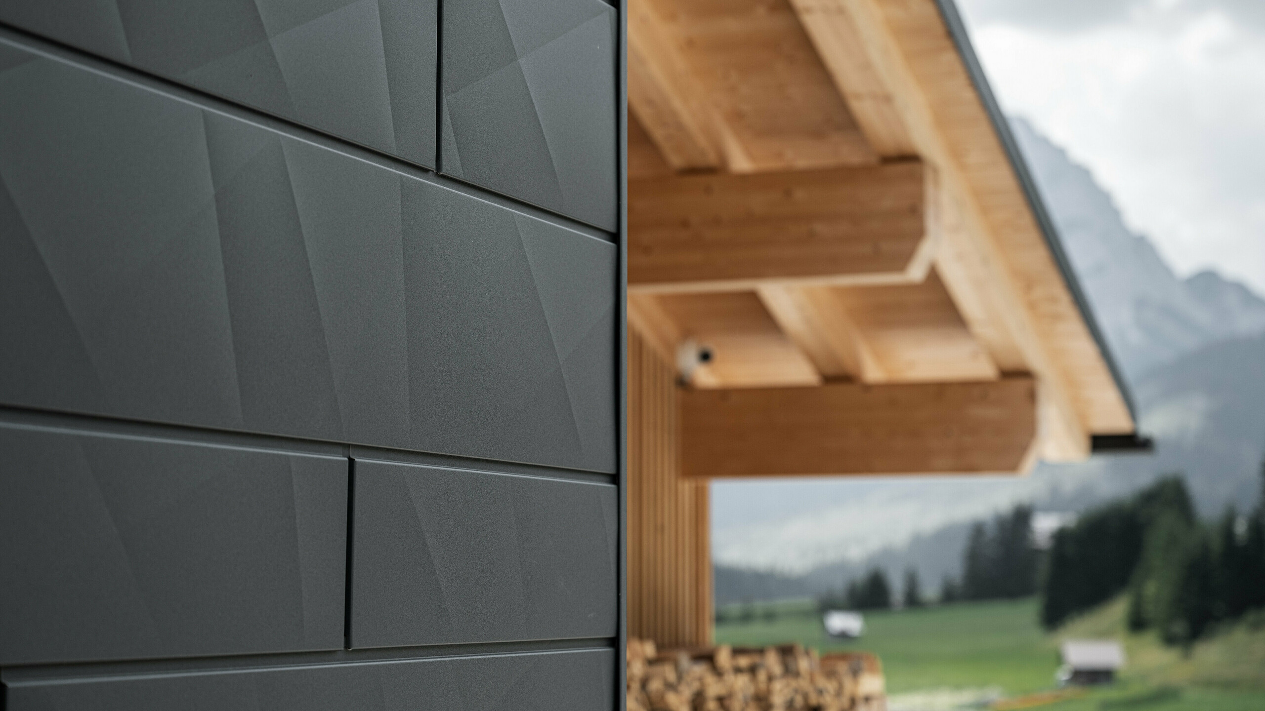 Nærbillede af Siding.X facadeplader i P.10 sort fra PREFA på Casa Hoffe i Sappada, Italien. Facadesystemet i aluminium er monteret i et moderne, abstrakt design, der danner en flot kontrast til de synlige træelementer i tagkonstruktionen. Den innovative facadeløsning passer perfekt ind i det alpine miljø og står for en kombination af funktionalitet, holdbarhed og æstetisk byggekultur.