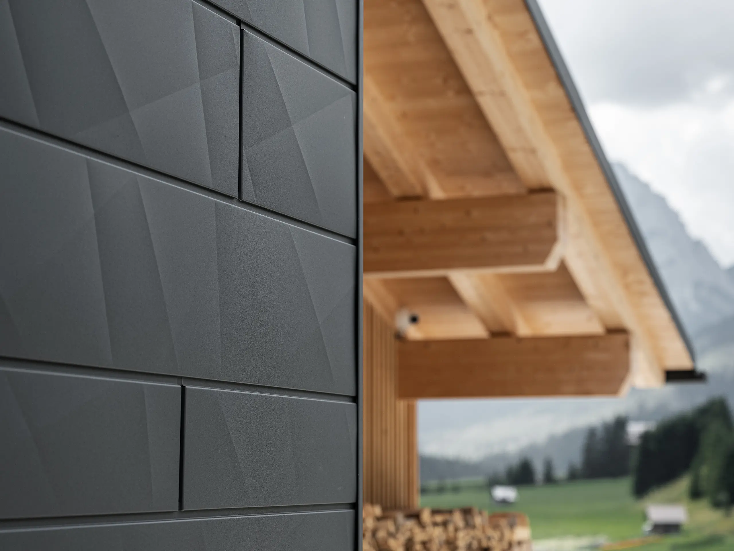 Nærbillede af Siding.X-facadepaneler i P.10 sort fra PREFA på Casa Hoffe i Sappada, Italien. Facadesystemet af aluminium er anbragt omhyggeligt i et moderne, abstrakt design, der giver en tilsvarende kontrast til de synlige træelementer i tagkonstruktionen. Den nyskabende facadeløsning føjer sig harmonisk ind i det alpine miljø og repræsenterer forbindelsen mellem funktioner, bestandighed og æstetisk byggekultur.