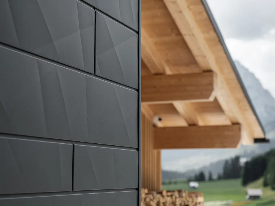 Nærbillede af Siding.X-facadepaneler i P.10 sort fra PREFA på Casa Hoffe i Sappada, Italien. Facadesystemet af aluminium er anbragt omhyggeligt i et moderne, abstrakt design, der giver en tilsvarende kontrast til de synlige træelementer i tagkonstruktionen. Den nyskabende facadeløsning føjer sig harmonisk ind i det alpine miljø og repræsenterer forbindelsen mellem funktioner, bestandighed og æstetisk byggekultur.