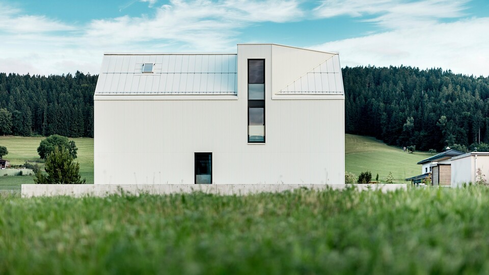 Das Bild zeigt das Einfamilienhaus in Freistadt aus einer Seitenansicht, die die markante, minimalistische Architektur des Gebäudes hervorhebt. Die Fassade aus reinweißem PREFALZ-Aluminium und das steile Satteldach schaffen eine klare, moderne Silhouette, die sich harmonisch in die grüne Umgebung einfügt. Die großen Fensterflächen und das elegante Gaubenfenster betonen das funktionale Design, während das hochwertige Aluminiummaterial sowohl ästhetische Beständigkeit als auch Witterungsresistenz gewährleistet. Das Gebäude strahlt durch seine einfache Form und die Verwendung langlebiger Materialien eine zeitlose Eleganz aus und stellt eine perfekte Kombination aus Architektur und Naturverbundenheit dar.