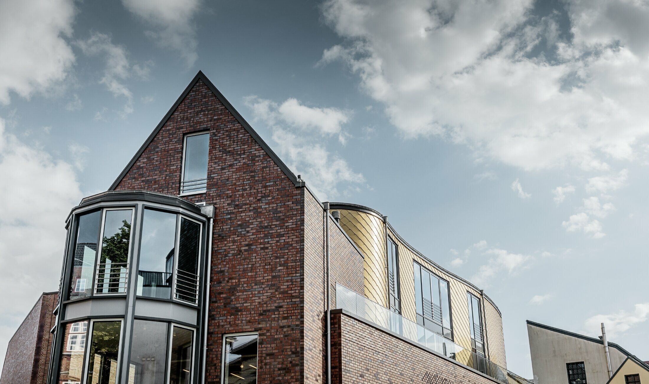 Erhvervsbygning med murstensfacade og store glaspartier. En del af den buede facade er beklædt med den gyldne vægrombe af aluminium fra PREFA.