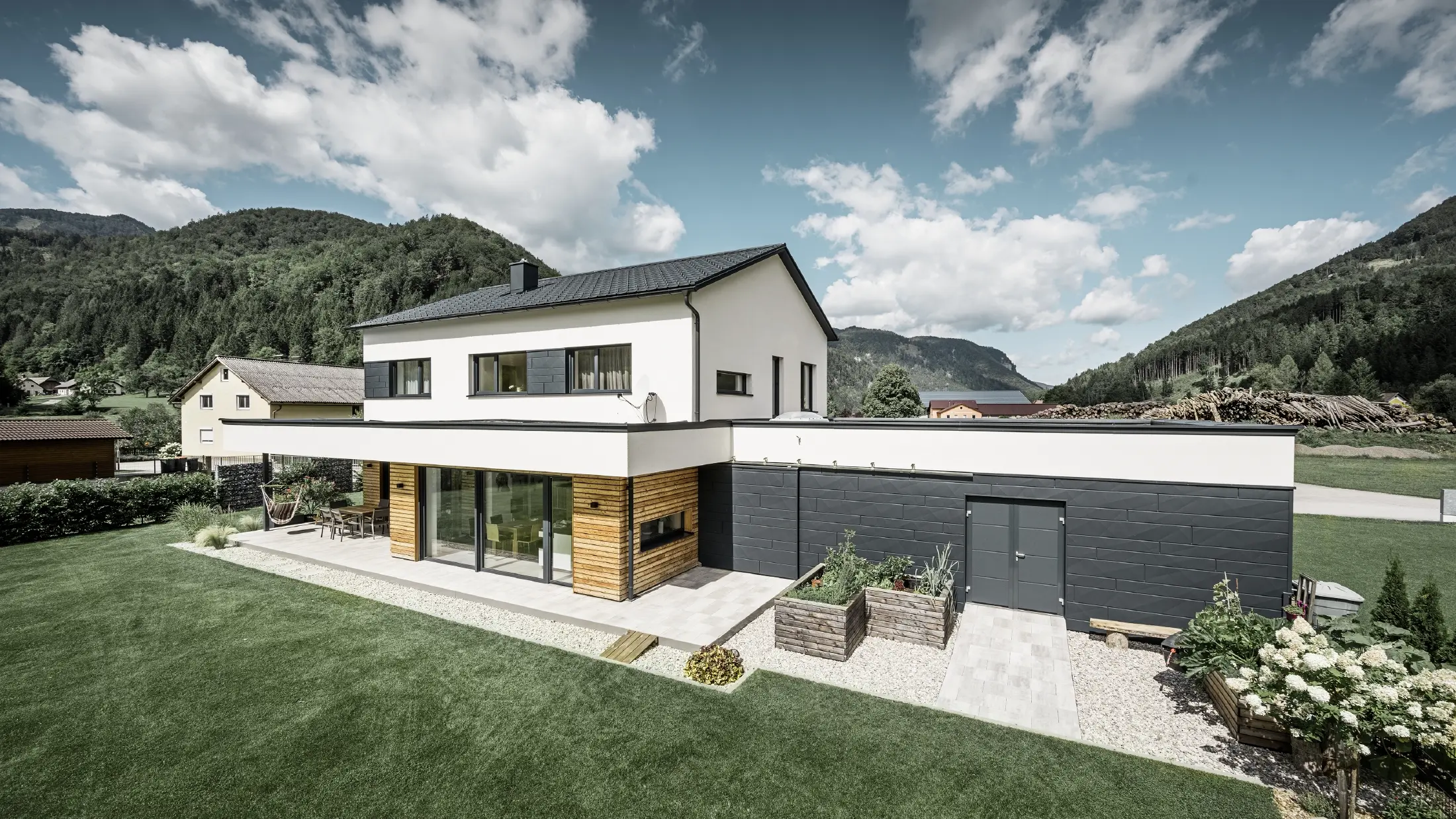 Moderne parcelhus med hvidpudset facade og PREFA Siding.X i P.10 antracit, som danner en markant kontrast til de naturlige omgivelser. Inddækket med PREFA tagplade, tagafvanding med PREFA hængende tagrende og nedløbsrør.
