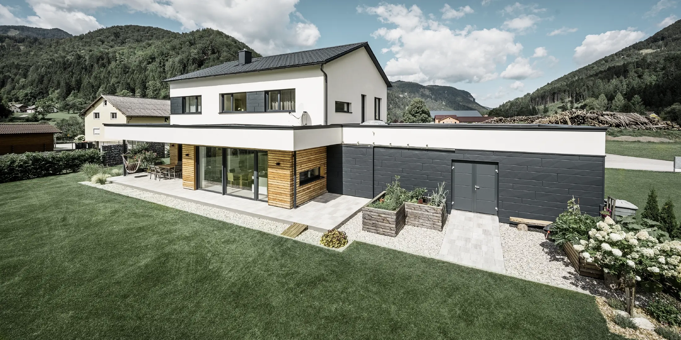 Moderne parcelhus med hvidpudset facade og PREFA Siding.X i P.10 antracit, som danner en markant kontrast til de naturlige omgivelser. Inddækket med PREFA tagplade, tagafvanding med PREFA hængende tagrende og nedløbsrør.