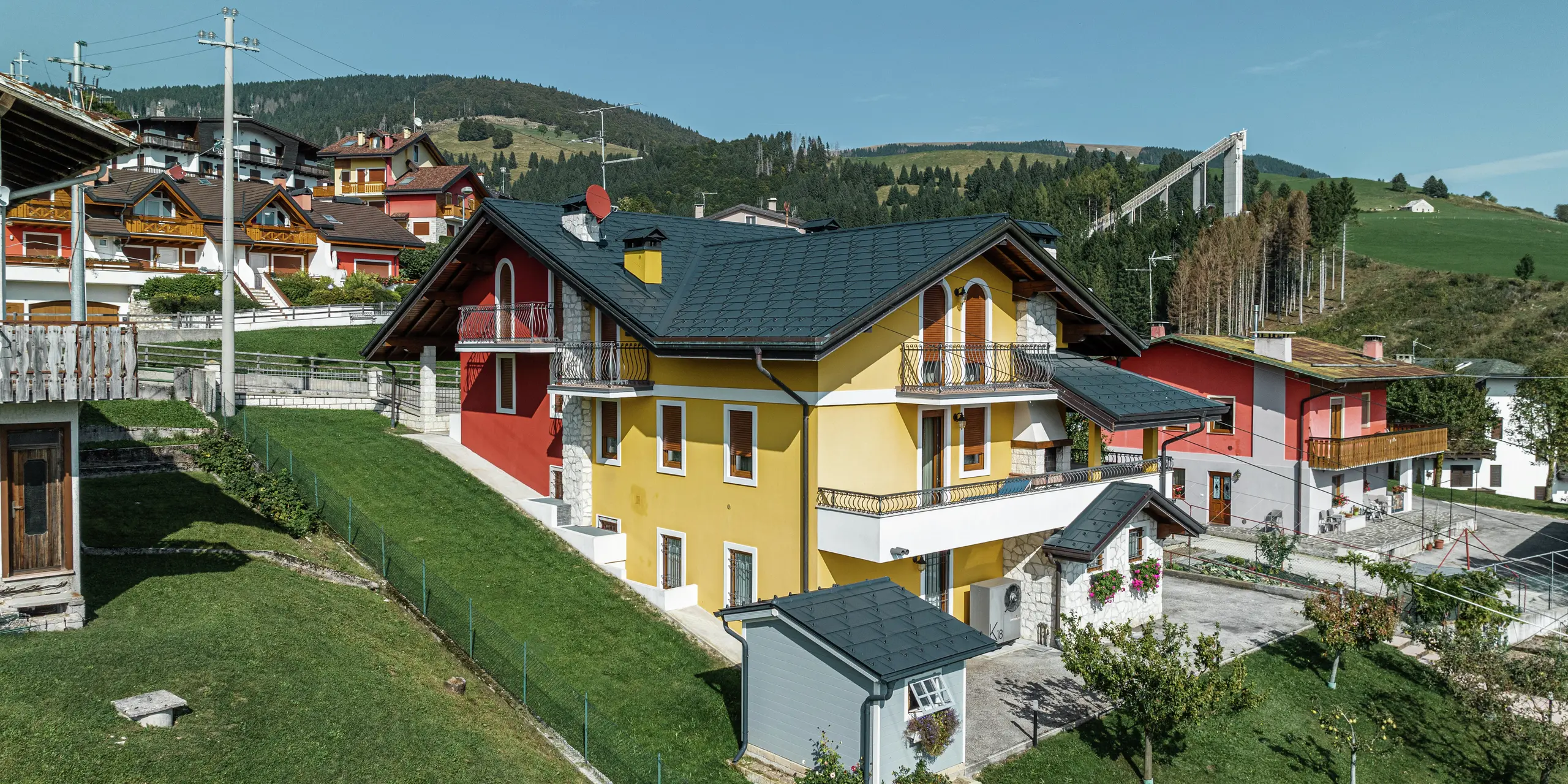 Einfamilienhaus in Gallio in traditioneller Alpenarchitektur mit leuchtend gelb-roter Putzfassade und Natursteinelementen. Das Dach aus PREFA Dachplatten R.16 in P.10 Dunkelgrau bietet höchste Witterungsbeständigkeit und harmoniert perfekt mit der Umgebung. Die Dachentwässerung mit PREFA Hängerinne und Ablaufrohr - ebenfalls in P.10 Dunkelgrau - sorgt für zuverlässigen Schutz. Umgeben von grünen Wiesen und mit Blick auf die Berge vereint das Haus Ästhetik, Langlebigkeit und Funktionalität. Im Hintergrund ist die bekannte Skisprungschanze der Region zu erkennen.