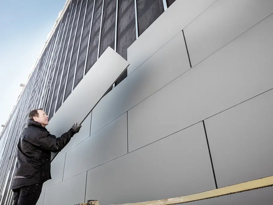 Fotos af en delvist færdig facade, der er beklædt med PREFA Sidings i bredde 500. Blikkenslageren monterer et facadepanel mere. Billedet er taget nedefra. 