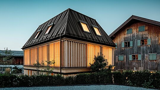 Das preisgekrönte „Badehaus“ des Hotel Hirschen in Schwarzenberg im stimmungsvollen Abendlicht. Im Vordergrund dominiert das moderne Walmdach mit markantem PREFA Stehfalz in P.10 Nussbraun – ein echtes architektonisches Statement, das durch seine unkonventionelle Linienführung und die 60-Grad-Neigung ins Auge sticht. Die Fassade aus vertikalen, geschoßhohen Holzlamellen ergänzt das historische Ensemble harmonisch und bringt modernen Minimalismus in Einklang mit der Bregenzerwälder Bautradition. Im Hintergrund ist das denkmalgeschützte Stammhaus sichtbar – ein kraftvoller Kontrast von Alt und Neu. Ein gelungenes Beispiel für zukunftsorientiertes Bauen mit hochwertigen PREFA Materialien.