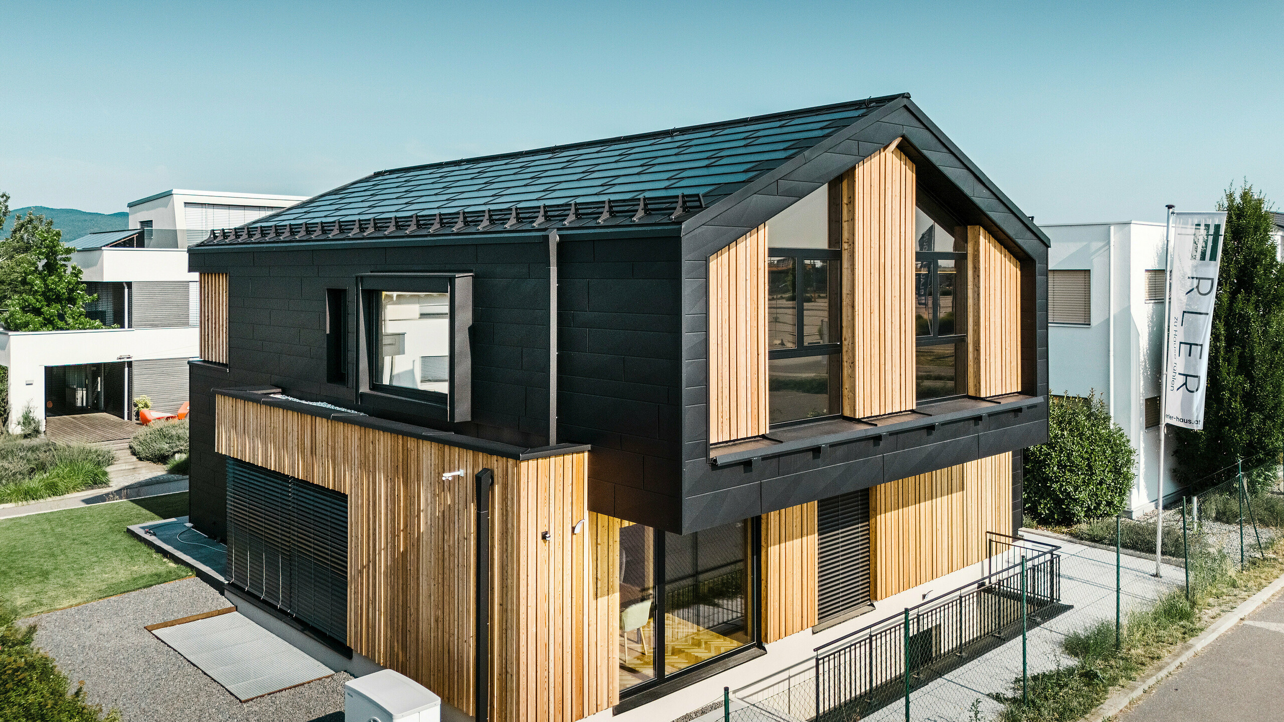 forside af modelhuset Autark i præfabrikationscenteret Blaue Lagune, Vösendorf (AT). Taget er dækket med sorte PREFA-solcelletagplader og tagplade R.16 i højkvalitets P.10-belægning. Facaden består af en kombination af lodrette træelementer og PREFA Siding.X i P.10 sort, hvilket giver bygningen et moderne og bæredygtigt udseende. I højre side af billedet vajer Erler Bau GmbH's flag.