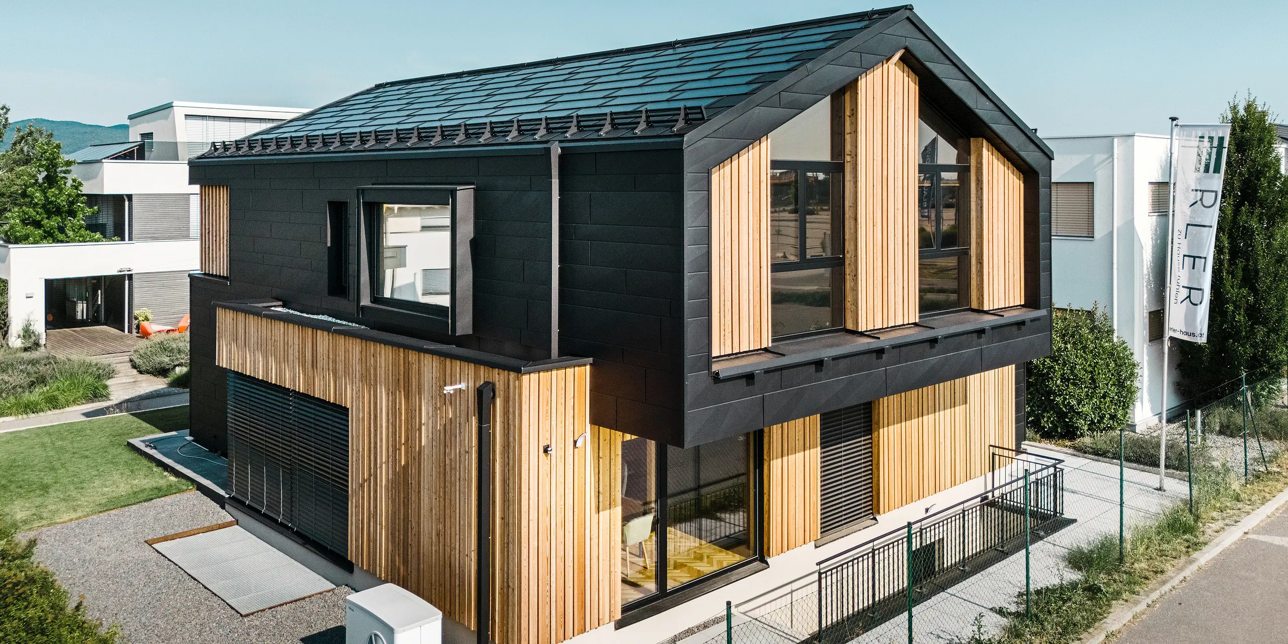 forside af modelhuset Autark i præfabrikationscenteret Blaue Lagune, Vösendorf (AT). Taget er dækket med sorte PREFA-solcelletagplader og tagplade R.16 i højkvalitets P.10-belægning. Facaden består af en kombination af lodrette træelementer og PREFA Siding.X i P.10 sort, hvilket giver bygningen et moderne og bæredygtigt udseende. I højre side af billedet vajer Erler Bau GmbH's flag.
