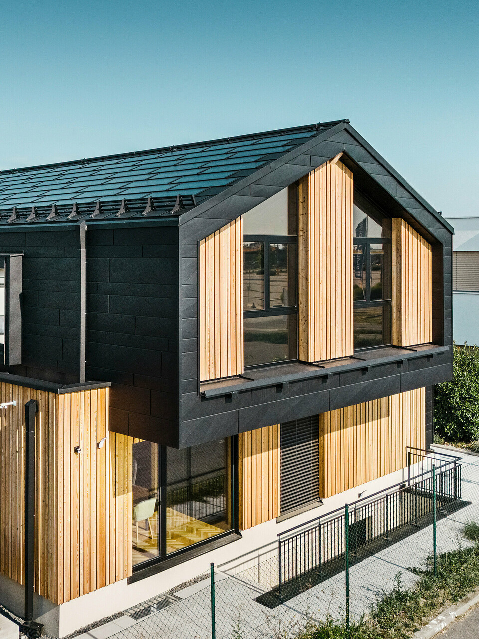 forside af modelhuset Autark i præfabrikationscenteret Blaue Lagune, Vösendorf (AT). Taget er dækket med sorte PREFA-solcelletagplader og tagplade R.16 i højkvalitets P.10-belægning. Facaden består af en kombination af lodrette træelementer og PREFA Siding.X i P.10 sort, hvilket giver bygningen et moderne og bæredygtigt udseende. I højre side af billedet vajer Erler Bau GmbH's flag.