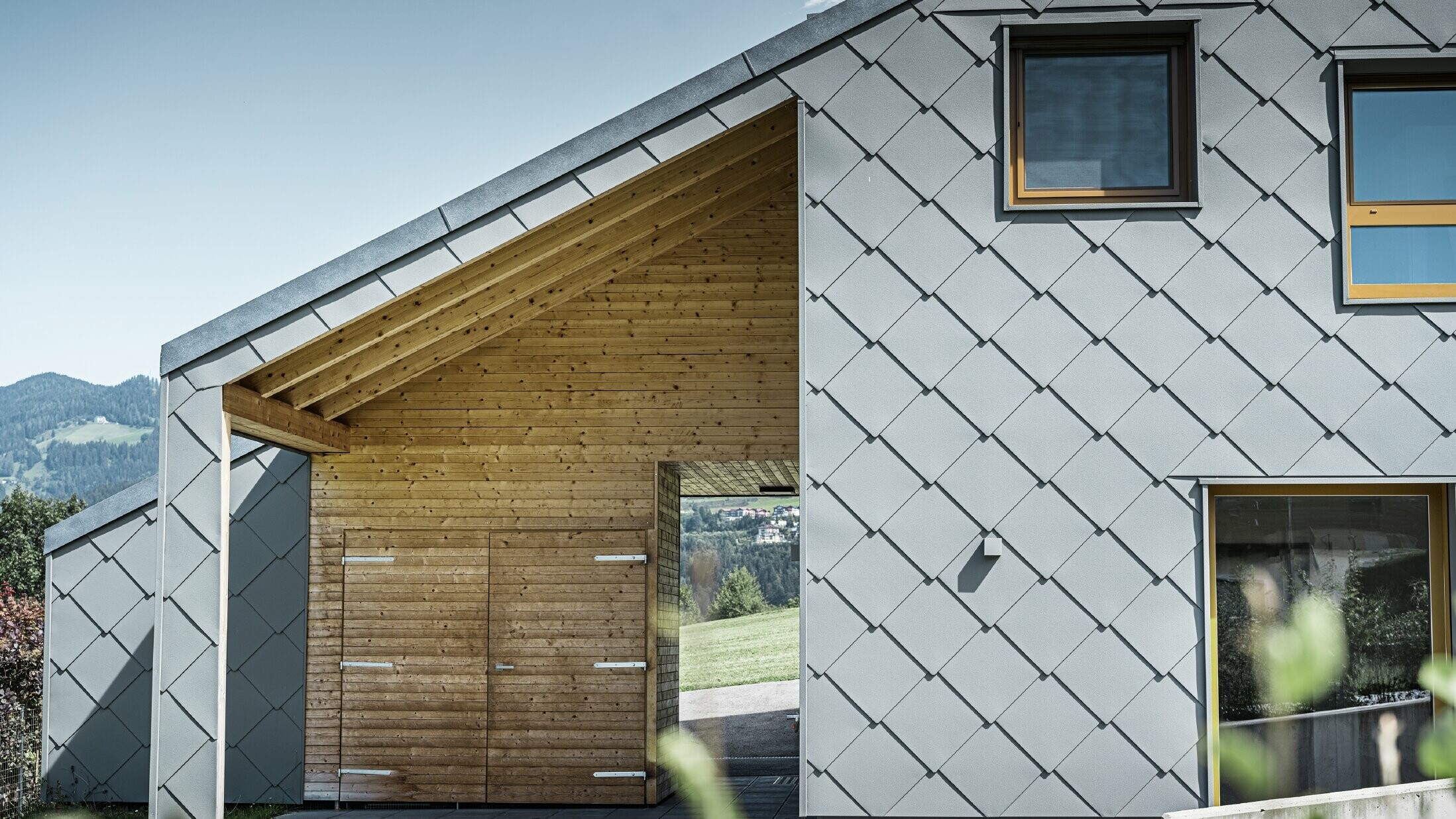 Det overdækkede indgangsområde er lavet med vandret træbeklædning, og resten af facaden er beklædt med den store PREFA-vægrombe af aluminium i lys grå