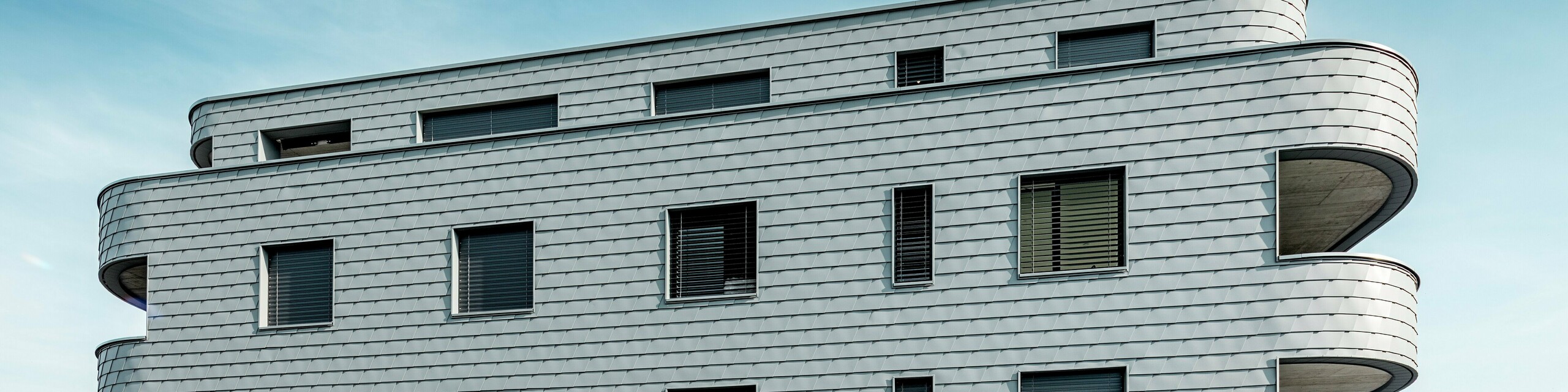 Frontalansicht des Bernerhauses in Buchs mit weich gerundeten Gebäudeecken und durchgehender Fassade aus PREFA Wandschindeln in P.10 Hellgrau. Die schuppenartig verlegte Aluminiumfassade erzeugt ein lebendiges Muster und betont die horizontale Linienführung der geschwungenen Balkonbänder. Präzise integrierte Fensteröffnungen fügen sich harmonisch in die homogene Oberfläche ein. Dank des leichten, witterungsbeständigen und langlebigen PREFA Aluminiums entsteht eine wartungsarme, formstabile Gebäudehülle mit markanter Ausstrahlung vor alpiner Kulisse.