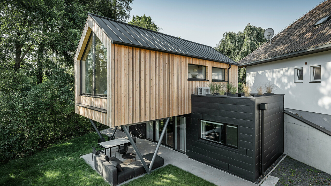 Das 'Woodstock Chalet 5' beeindruckt mit einer eindrucksvollen Kombination aus natürlichen Holzoberflächen und den modernen PREFA Siding.X Fassadenelementen in Anthrazit. Die überhängende Holzstruktur des Obergeschosses ragt markant über die dunklen Fassadenelemente, die das Erdgeschoss verkleiden, und erzeugt so ein Spiel aus Licht und Schatten. Die klaren Linien und die robuste Konstruktion der Siding.X Elemente ergänzen das innovative Design des Gebäudes und bieten einen spannenden visuellen Kontrast zur umgebenden Vegetation und dem traditionellen Gebäude im Hintergrund. Am Dach des Chalets wurde das PREFALZ Dachsystem in Form einer gleichmäßigen Stehfalzdeckung verlegt. Am rechten Rand des Erdgeschosses ist das PREFA Dachentwässerungssystem zu erkennen, das analog zur Aluminiumfassade in Anthrazit gehalten ist. Für die Dachentwässerung wurden der Wasserfangkasten gefolgt vom Ablaufrohr montiert.