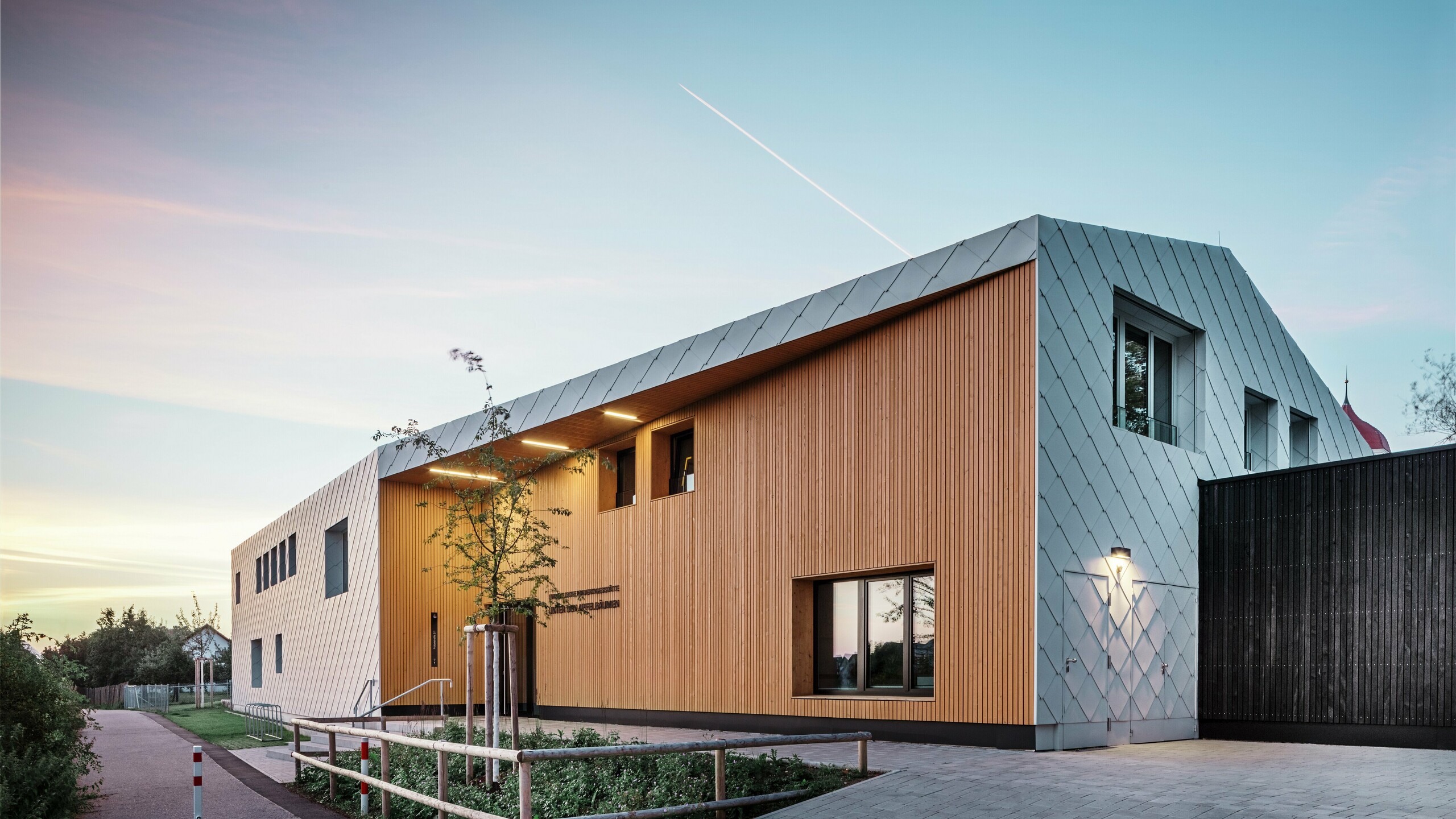 Foto taget i skumringen af en moderne daginstitution i Ulm. Taget blev designet uden udhæng. Størstedelen af facaden er beklædt med store aluminiumsromber fra PREFA i hvid, og indgangspartiet er i stor udstrækning beklædt med lodrette træpaneler.