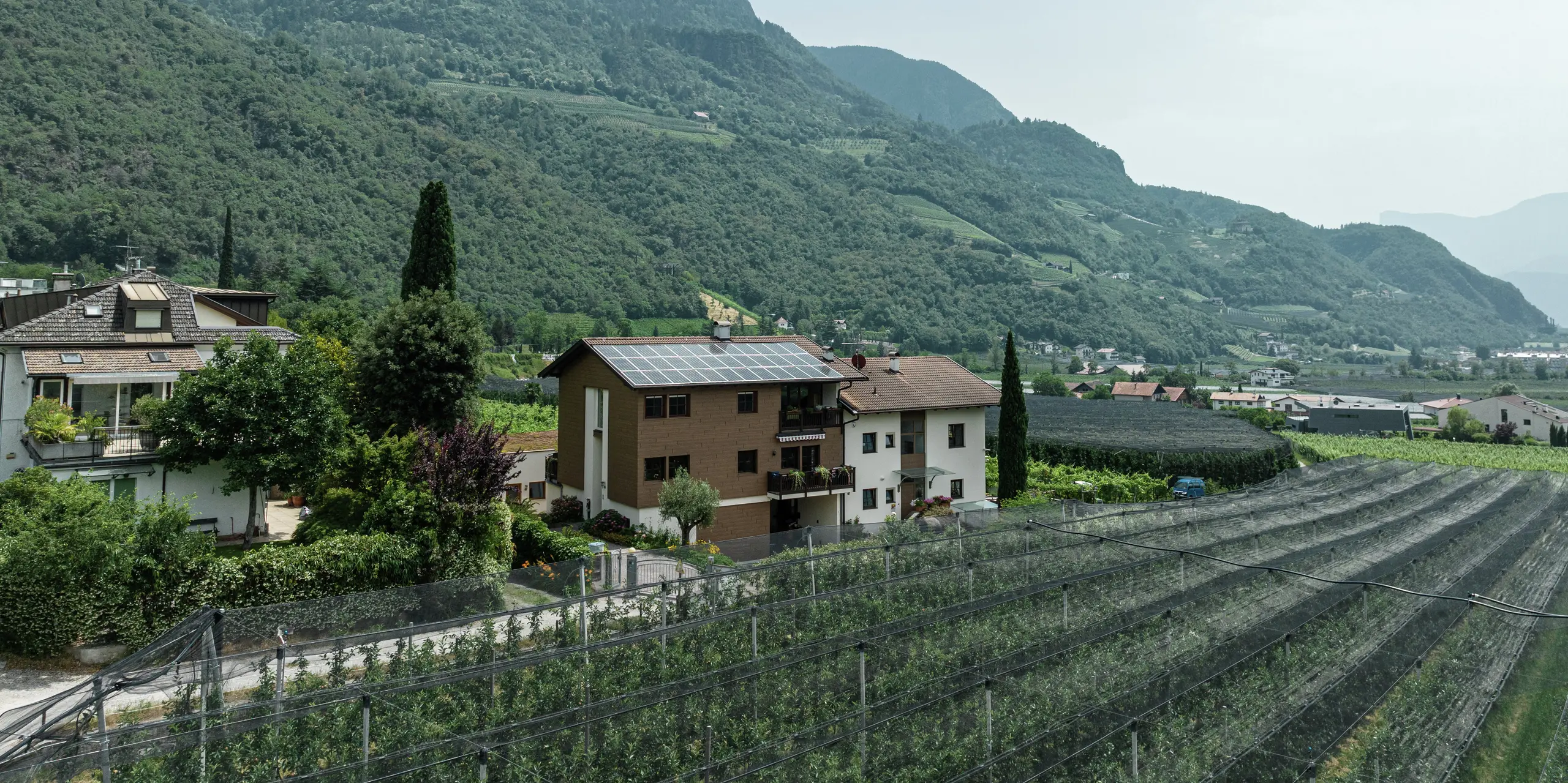 Luftaufnahme der Ferienwohnungen Prechtlhof in Meran, Südtirol, umgeben von Obstgärten und Berglandschaft. Die modernisierte Fassade mit PREFA Sidings in Wallnussbraun sorgt für eine natürliche Holzoptik aus langlebigen, robusten und wartungsarmen Aluminium. Die Photovoltaikanlage auf dem Dach trägt zur nachhaltigen Energieversorgung bei.