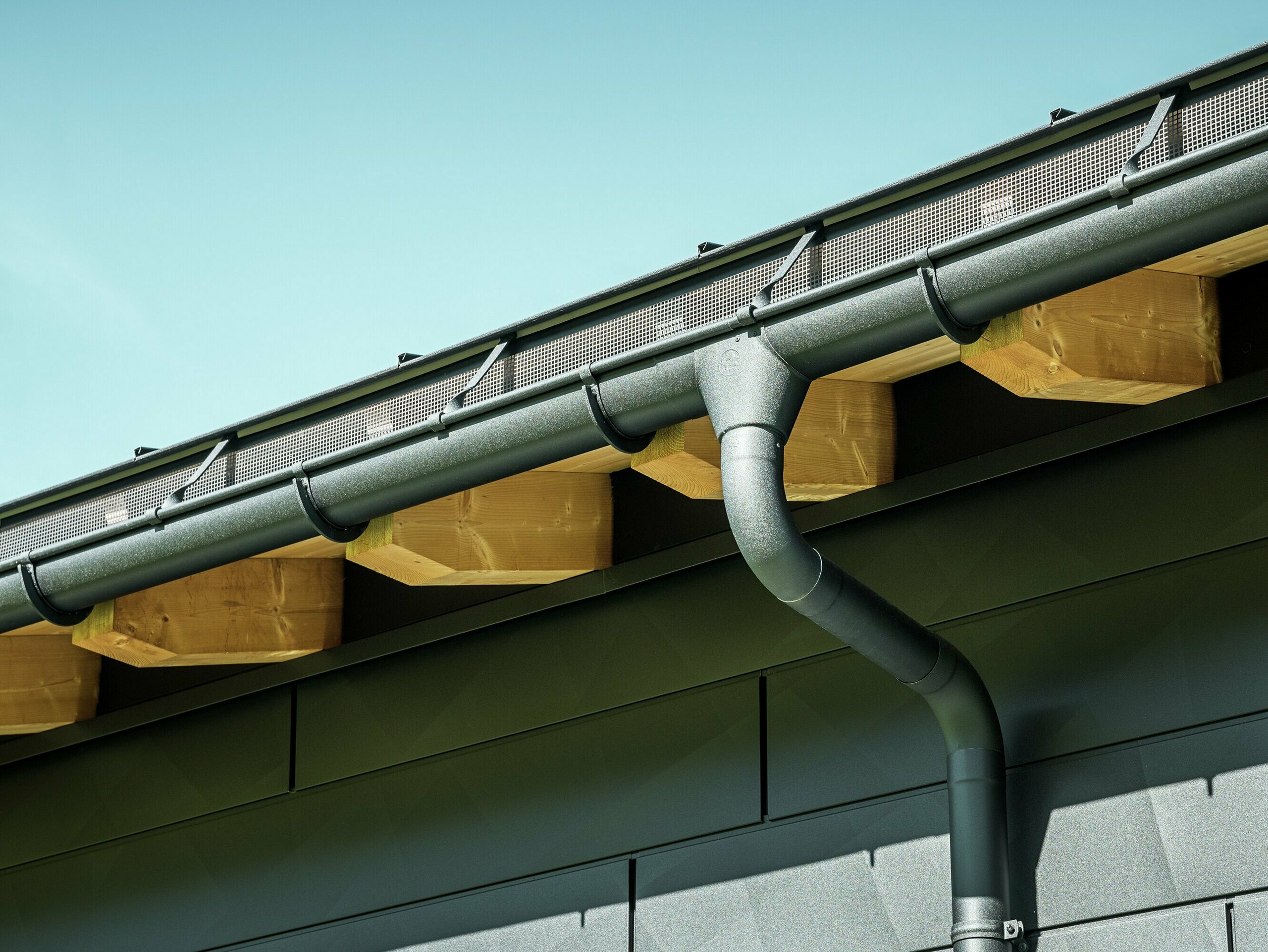 Nærbillede af facaden på et moderne enfamiliehus i Ascharina, Schweiz. Bygningens sider er beklædt med de markante PREFA Siding.X facadepaneler i P.10 sort. PREFA tagafvanding med halvrund tagrende og nedløbsrør er farveafstemt.