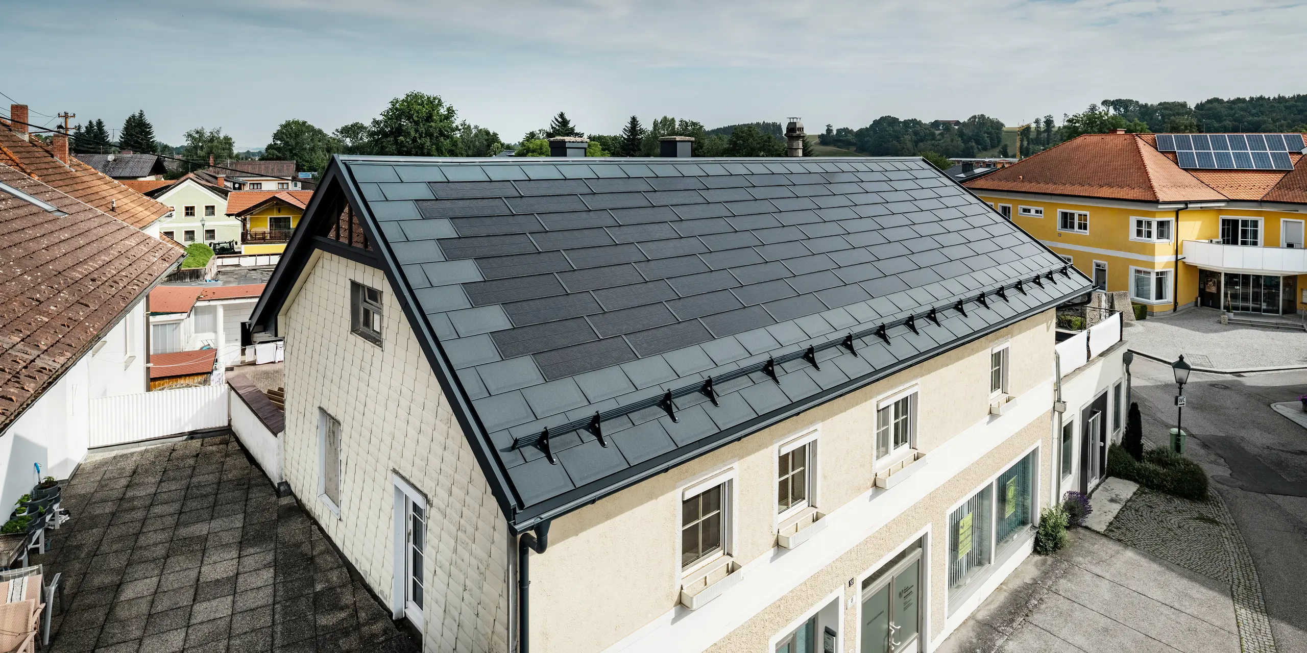 Udsigt fra oven over et parcelhus i Mettmach, Østrig. Den  ældre bygning blev renoveret med antracitfarvede PREFA-solcelletagplader. Snebeskyttelsessystemerne og solcelletagpladernes klare linjer understreger det rene håndværk i PREFAs aluminiumsprodukter. Det orangefarvede tegltag på nabobygningen står i kontrast i baggrunden. Solcellemodulerne er installeret på taget af det nærliggende parcelhus i en klassisk tagløsning, der viser mangfoldigheden i energieffektivt tagdesign.