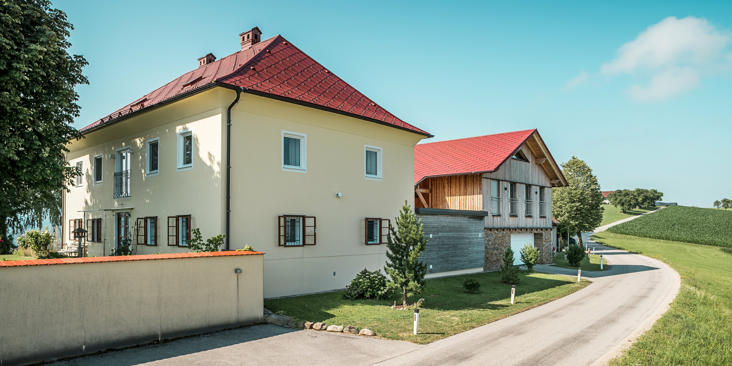 Blick von der Straße auf die sanierten Einfamilienhäuser in Aschach an der Steyr. Die PREFA Dachraute 44 x 44 in der Farbe P.10 Oxydrot sticht in der ländlichen Umgebung hervor. Die markante rote Farbe der Aluminiumdächer bildet einen harmonischen Kontrast zur modernen Fassade und zum traditionellen Holzbau des Nebengebäudes. Die PREFA-Produkte sind nicht nur eine ästhetische Bereicherung, sondern überzeugen auch durch ihre Beständigkeit und Langlebigkeit gegenüber den Witterungseinflüssen der Region.