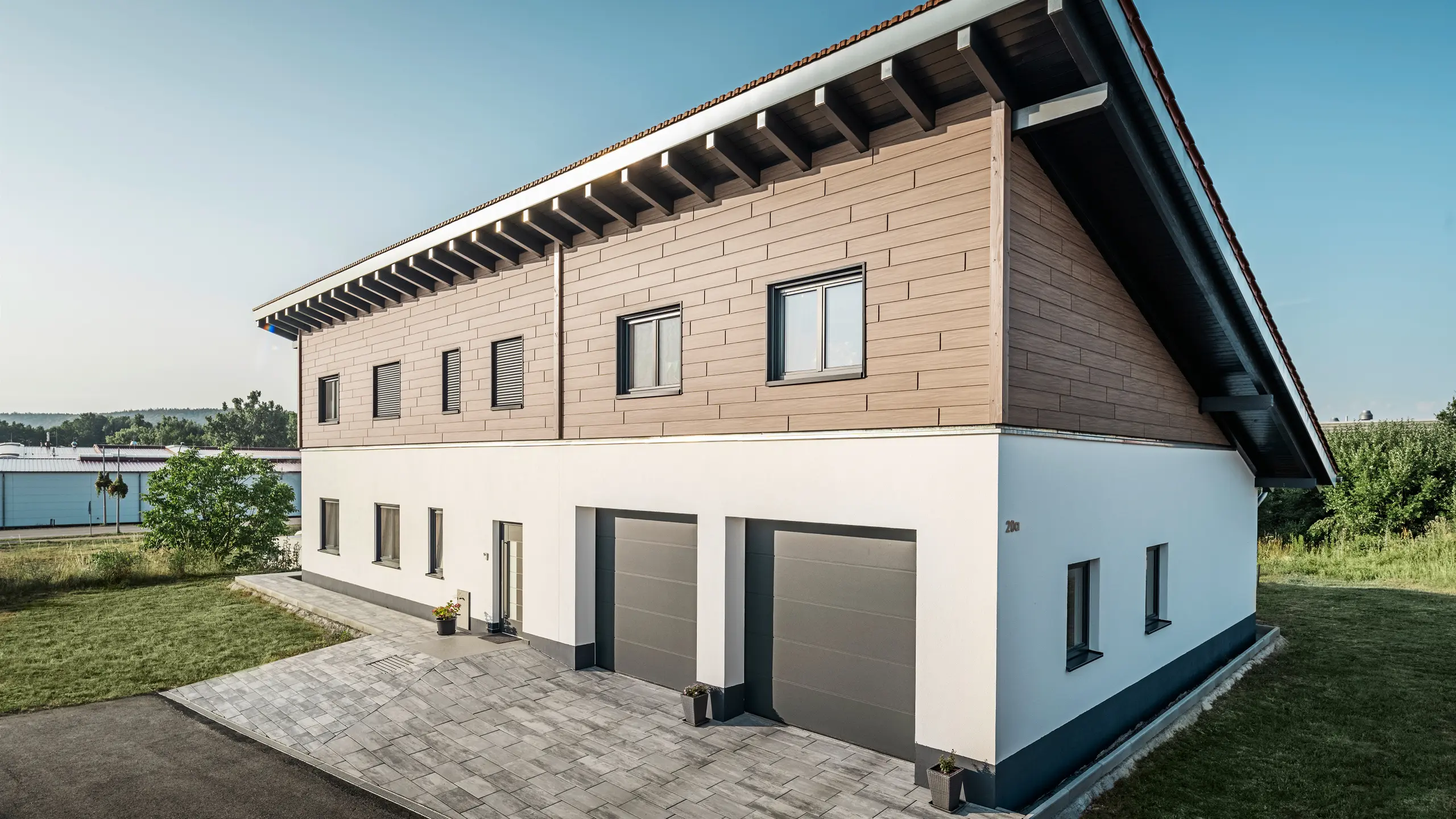 Vy från sidan av en villa med pulpettak i Bruck. Det övre våningsplanet är klätt med PREFA Sidings i valnød. Till höger i bild syns två garageportar i antracit. Entrédörren är placerad i mitten av byggnaden. Sockeln är i en slät vit kulör. 