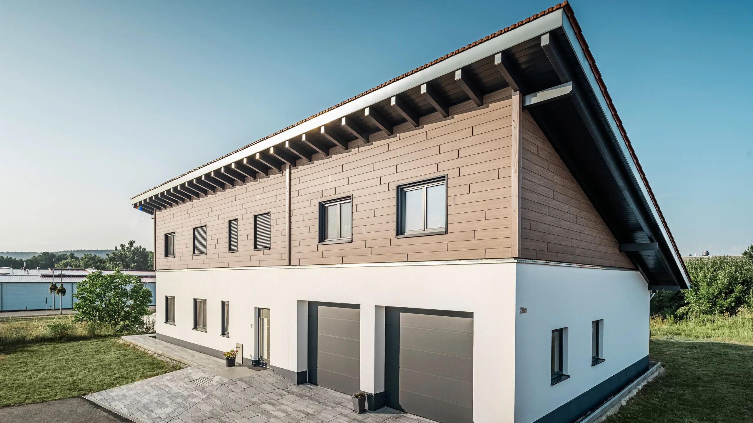Vy från sidan av en villa med pulpettak i Bruck. Det övre våningsplanet är klätt med PREFA Sidings i valnød. Till höger i bild syns två garageportar i antracit. Entrédörren är placerad i mitten av byggnaden. Sockeln är i en slät vit kulör. 