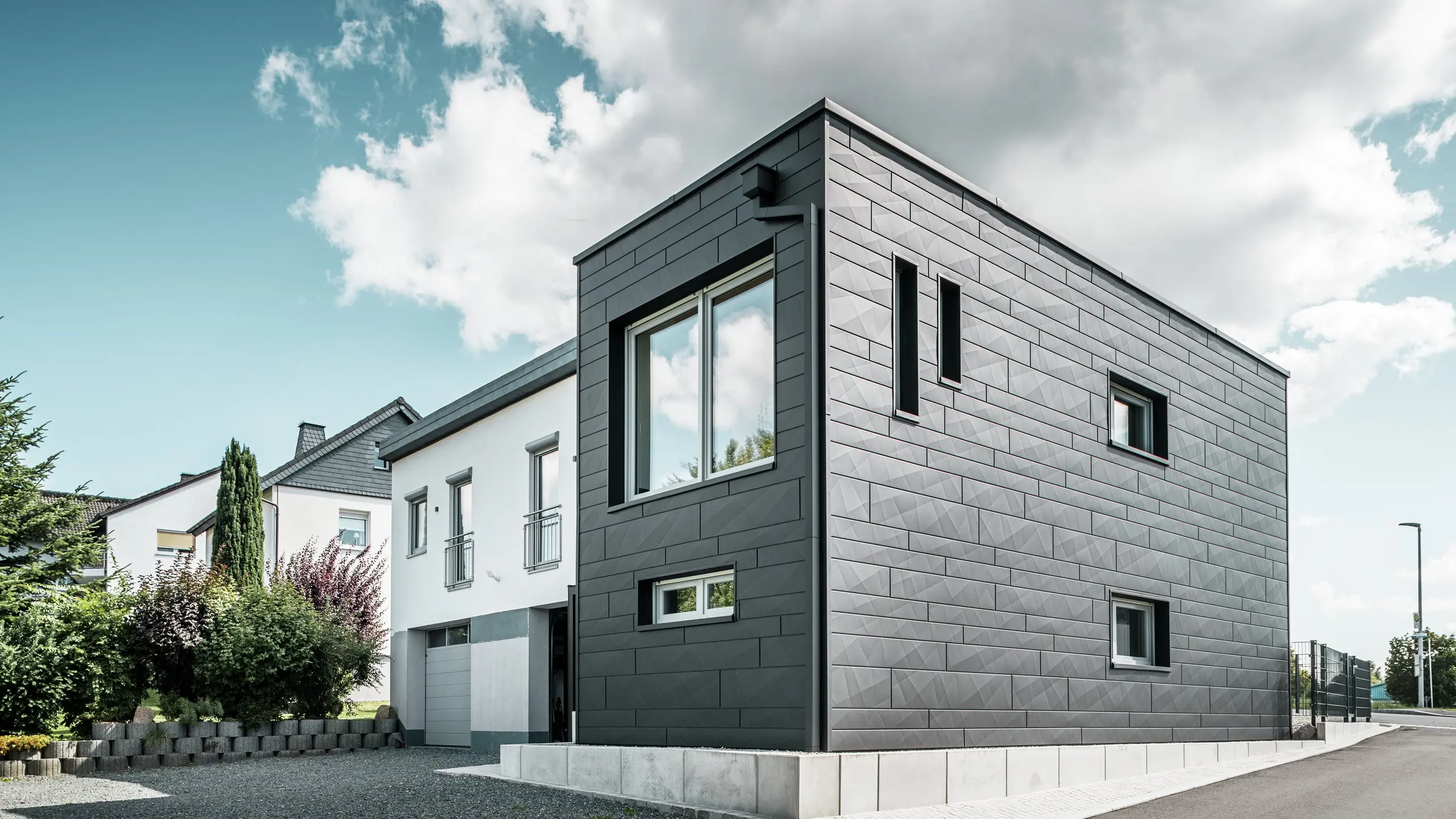 Den stilfulde og moderne facade på det renoverede enfamiliehus i Teuschnitz viser de klart strukturerede PREFA Siding.X-paneler i P.10 sort. Den geometriske placering af de højkvalitative pladepaneler giver bygningen en markant, tidløs æstetik. Vinduerne er indrammet af sorte aluminiumskompositplader, hvilket afrunder designet harmonisk. Det integrerede PREFA tagafvanding med kvadratrør sikrer en funktionel og æstetisk løsning.