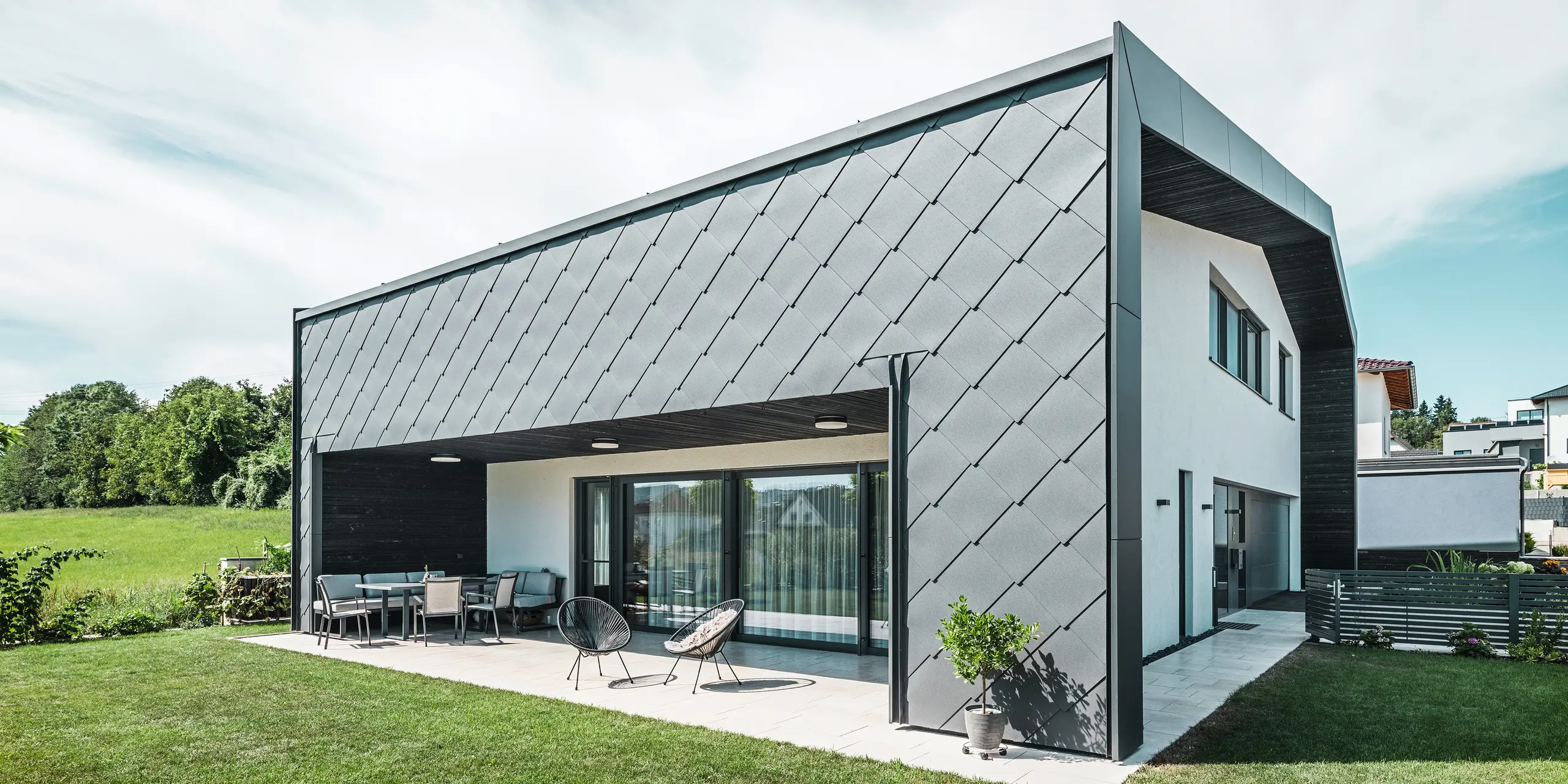 Velplejet haveområde til et enfamiliehus i Aschach. Bygningen er stilfuldt beklædt med mørk grå PREFA aluminium vægromber 44 × 44. Den overdækkede terrasse tilbyder et hyggeligt siddeområde med udsigt til den saftige grønne græsplæne. Den elegante aluminiumsbeklædning i kombination med hvide pudsflader sikrer en moderne og vejrbestandig arkitektur. Store vinduesflader lukker masser af dagslys ind i rummet og skaber en åben, lys atmosfære.