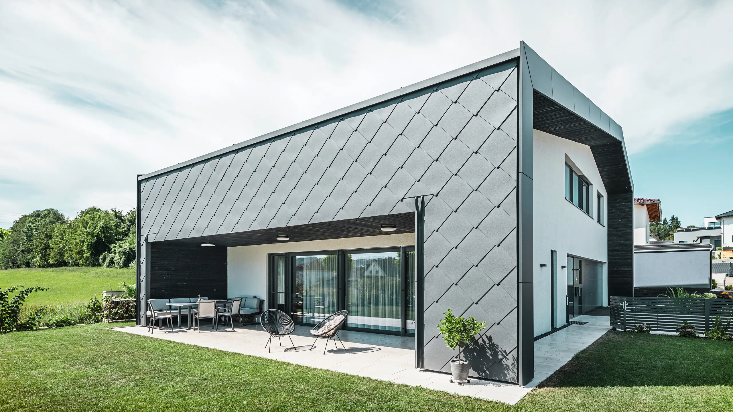 Velplejet haveområde til et enfamiliehus i Aschach. Bygningen er stilfuldt beklædt med mørk grå PREFA aluminium vægromber 44 × 44. Den overdækkede terrasse tilbyder et hyggeligt siddeområde med udsigt til den saftige grønne græsplæne. Den elegante aluminiumsbeklædning i kombination med hvide pudsflader sikrer en moderne og vejrbestandig arkitektur. Store vinduesflader lukker masser af dagslys ind i rummet og skaber en åben, lys atmosfære.
