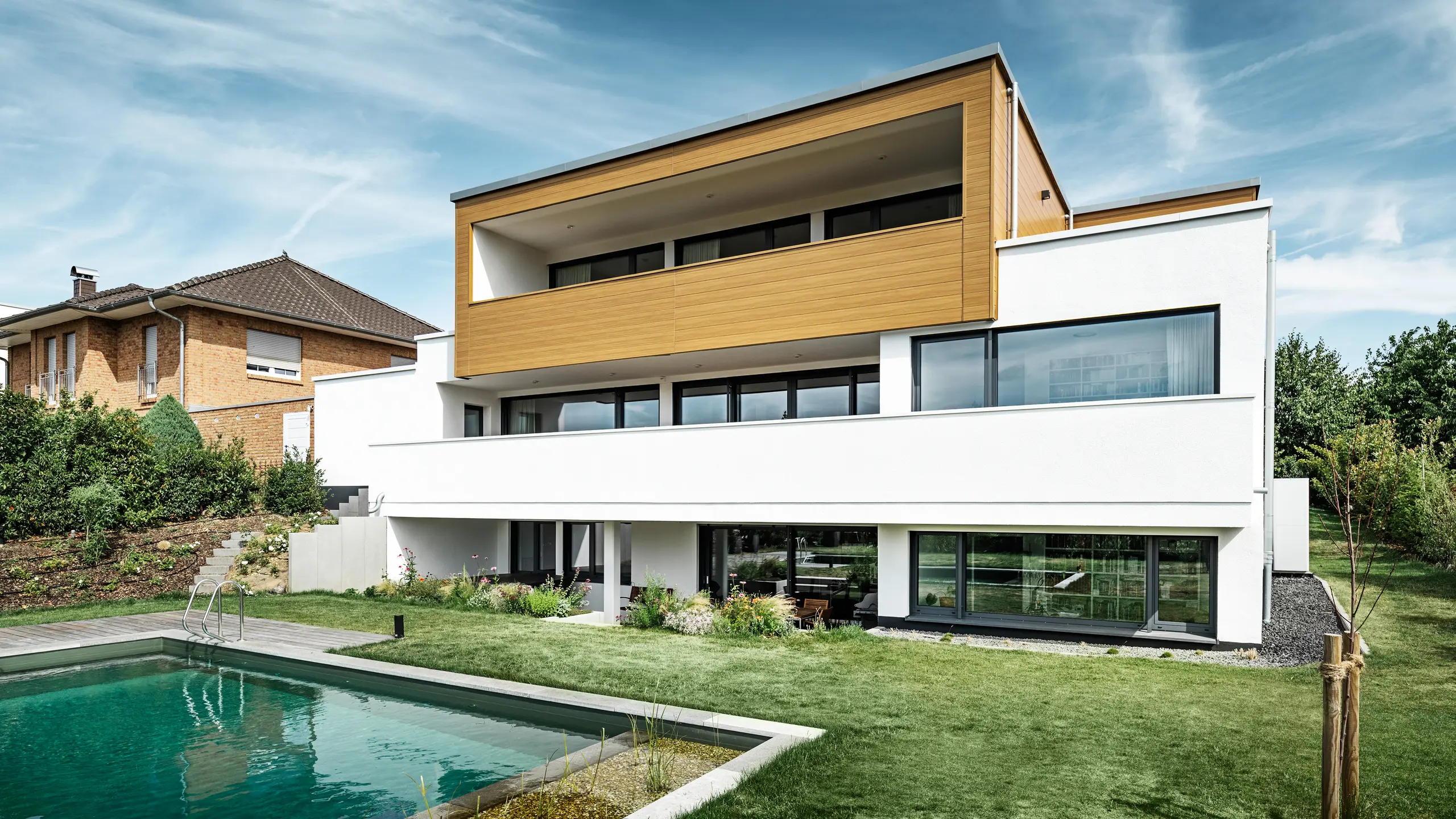 Baksidan av en modern villa i Usingen, Tyskland. Den gröna gräsmattan, en pool och ett ungt trädgårdsträd syns längst ner i bilden. Det iögonfallande huset har en vit byggnadsgrund av en putsad fasad, en aluminiumfasad på övervåningen av PREFA Sidings i natur eg och stora fönsterpartier med antracitfärgad karm. På avstånd syns grannhuset med tegelfasad i bildens vänstra kant.