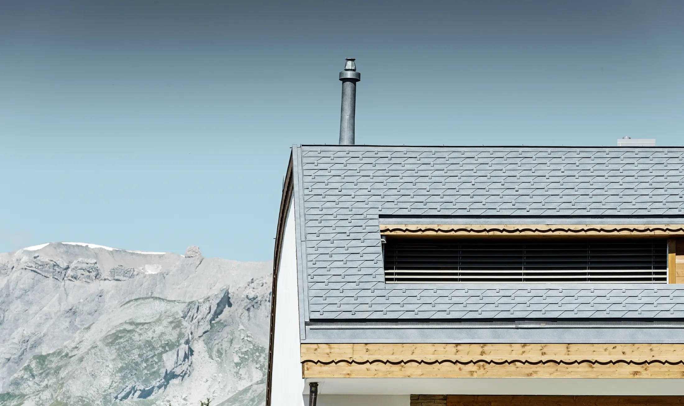 Flerfamiliehus i Crans-Montana med bjerge i baggrunden og en facade med fantasifulde træelementer med en PREFA aluminium tagspån-tag i stengrå