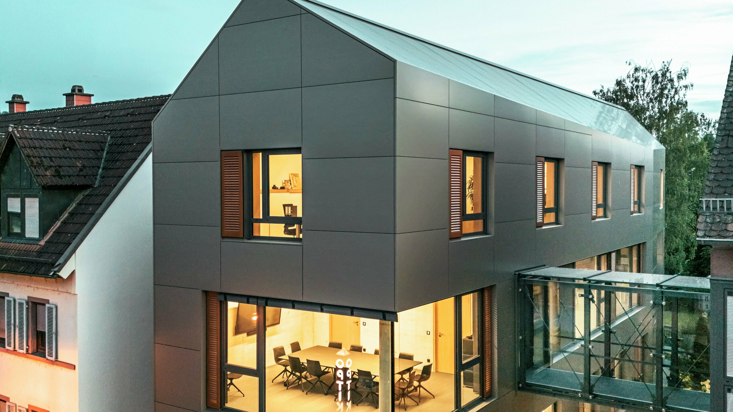 Slika prikazuje gradsku vijećnicu u Alzenauu u sumrak, s modernom fasadom od aluminijskih kompozitnih ploča PREFA u elegantnoj brončanoj boji. Stakleni spojni hodnik koji povezuje zgradu s obližnjim dijelom vizualno se ističe. Veliki prozori otkrivaju unutrašnjost, obasjanu toplim svjetlom. Jasni geometrijski oblik fasade i okolna zelena površina pojačavaju moderan, suvremen izgled zgrade.