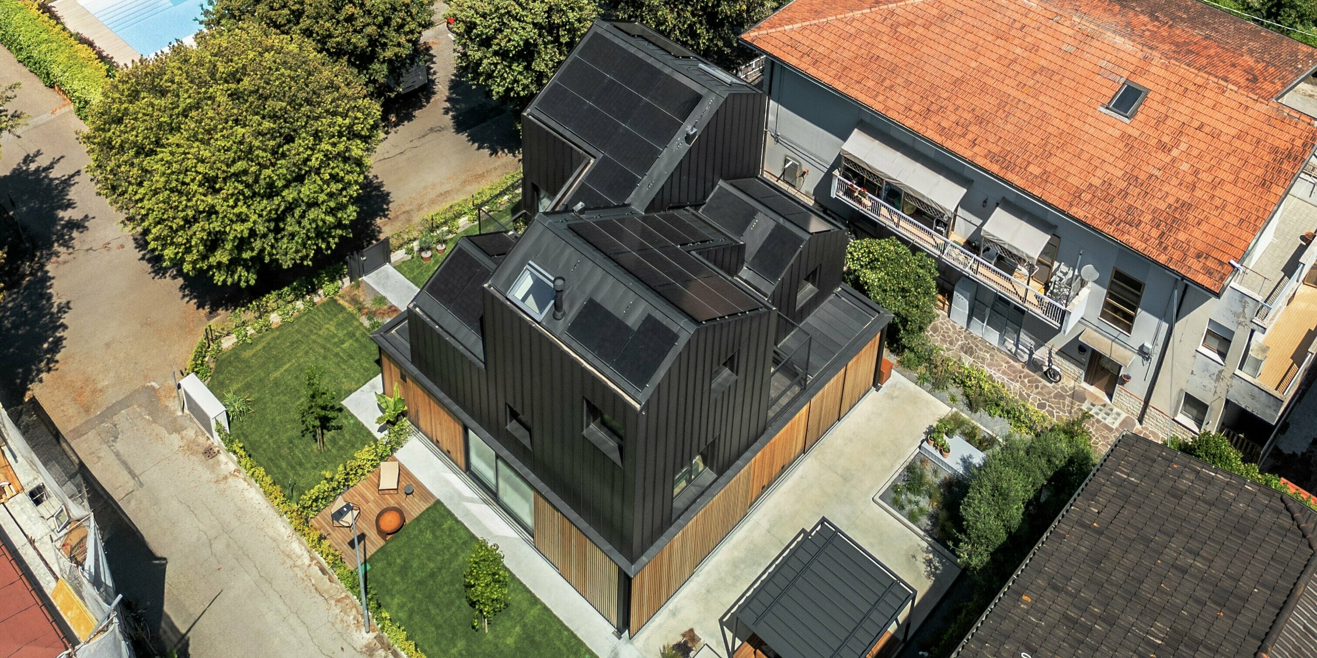 Luftaufnahme eines modernen Doppelhauses in Cattolica: markante Aluminiumfassade und Dach mit PREFALZ in Schwarzgrau, kombiniert mit Holzlamellen. Das Gebäude hebt sich durch klare Linien und hochwertige PREFA Materialien inmitten traditioneller Häuser mit roten Ziegeldächern und mediterraner Umgebung hervor.