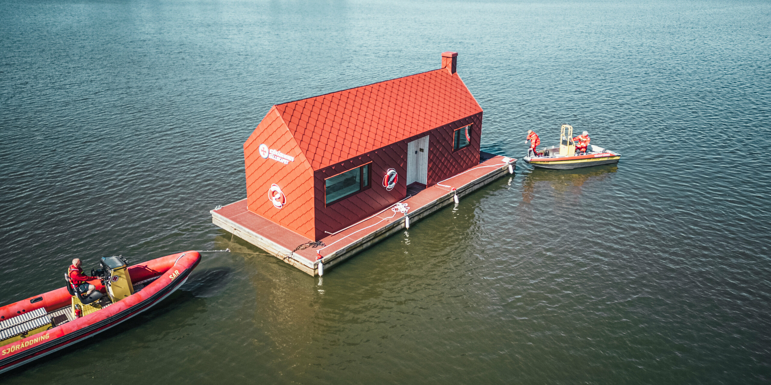 Das Seenotrettungsstationshaus auf dem Hjälmaren See in Schweden, erfasst aus der Vogelperspektive, hebt sich mit seiner oxydroten PREFA Aluminiumverkleidung deutlich von der umgebenden blauen Wasserfläche ab. Mit Dach- und Wandrauten 29×29 bietet es eine ästhetische und funktionale Struktur, die speziell für die maritimen Herausforderungen konzipiert wurde. Ein Rettungsboot liegt an der Anlegestelle bereit, während das Team Ausrüstung vorbereitet, ein stilles Zeugnis der ständigen Einsatzbereitschaft der Seenotretter in dieser friedlichen, aber potentiell dynamischen Umgebung.