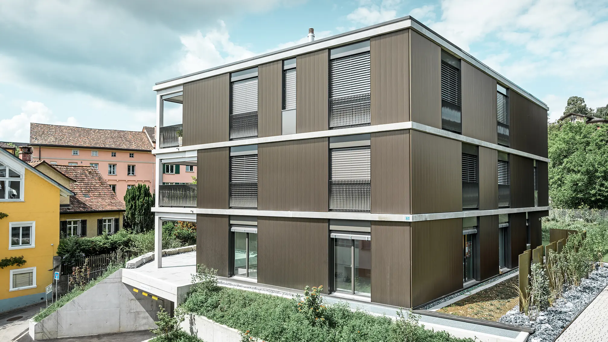 Den markante aluminiumsfacade på denne flerfamiliebolig i Schaffhausen er udstyret med det elegante PREFA takprofiler i farven Colinal. Den lodrette placering af de ekstruderede aluminiumsprofiler får bygningen til at virke mere kompakt, og facadestrukturen får en harmonisk balance. Det elegante design passer smagfuldt ind i omgivelserne og giver bygningen en moderne elegance, der både er visuelt tiltalende og funktionel. Perspektivet viser flerfamiliehuset fra forsiden med indkørslen til garagen. I baggrunden ses træer, andre bygninger og et kirketårn.