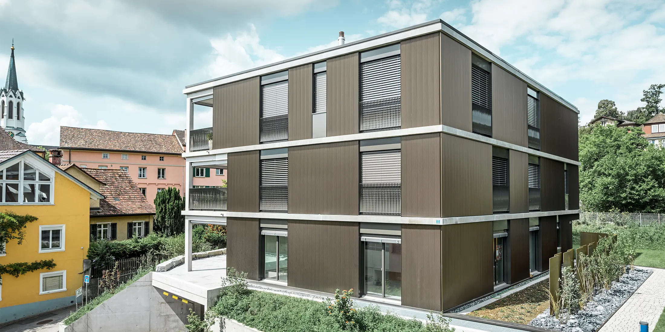 Den markante aluminiumsfacade på denne flerfamiliebolig i Schaffhausen er udstyret med det elegante PREFA takprofiler i farven Colinal. Den lodrette placering af de ekstruderede aluminiumsprofiler får bygningen til at virke mere kompakt, og facadestrukturen får en harmonisk balance. Det elegante design passer smagfuldt ind i omgivelserne og giver bygningen en moderne elegance, der både er visuelt tiltalende og funktionel. Perspektivet viser flerfamiliehuset fra forsiden med indkørslen til garagen. I baggrunden ses træer, andre bygninger og et kirketårn.