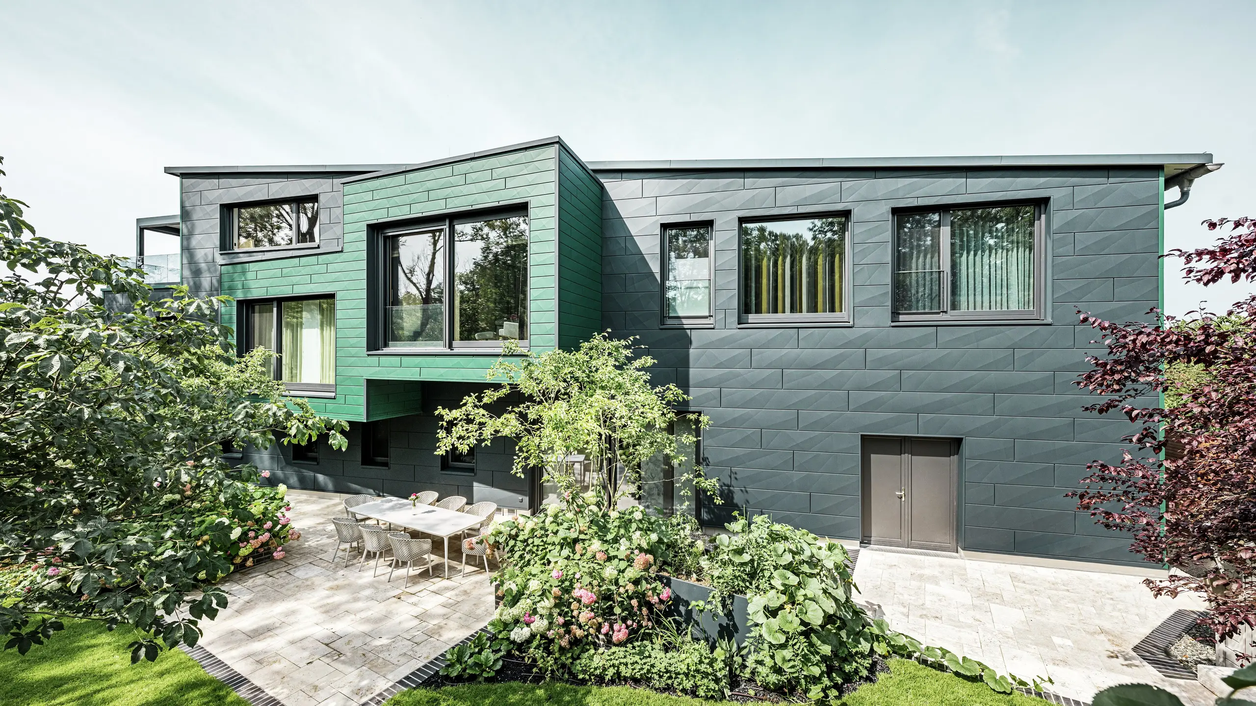 Enkeltstående hus i Türkheim imponerer med sin ekstraordinære facade, der er beklædt med PREFA Siding X-paneler i farverne antracit og mosgrøn. De klare linjer og det markante farvevalg på aluminiumpanelerne giver bygningen et moderne og elegant udtryk. I forgrunden ses den store have med en stilfuld terrasse og et spisebord med moderne stole. Farverige blomsterbede og sunde planter skaber en livlig, grøn kontrast til den robuste facade. Den holdbare og vedligeholdelsesvenlige aluminiumsfacade beskytter bygningen og sikrer tidløs skønhed.