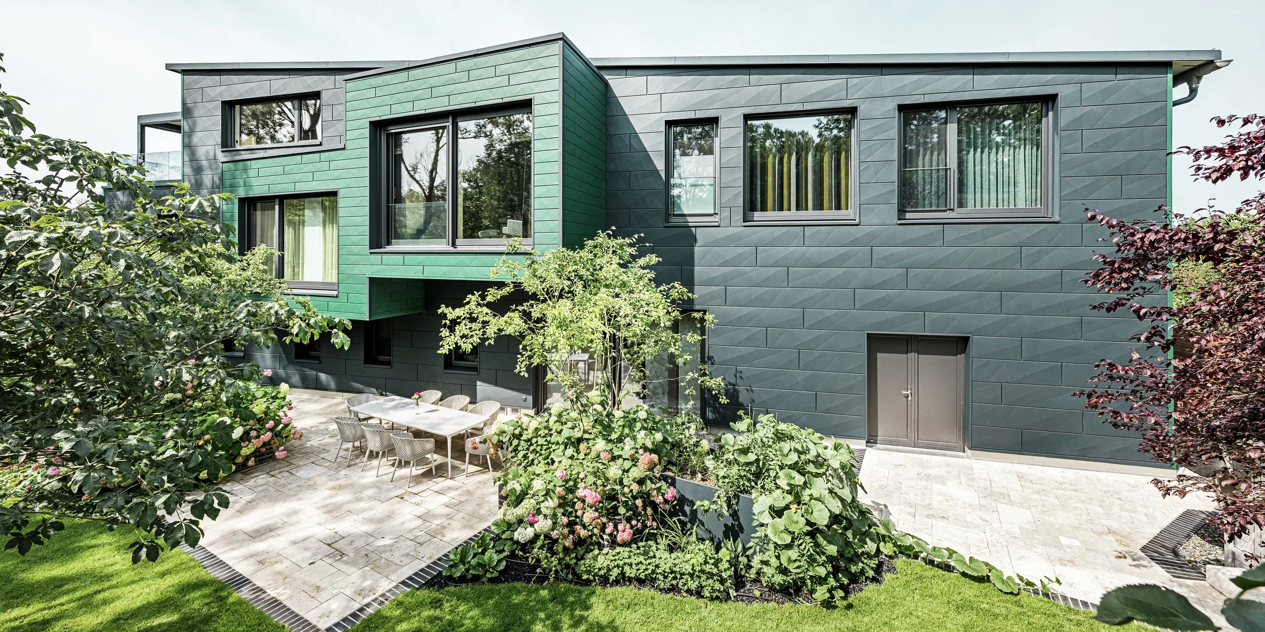 Enkeltstående hus i Türkheim imponerer med sin ekstraordinære facade, der er beklædt med PREFA Siding X-paneler i farverne antracit og mosgrøn. De klare linjer og det markante farvevalg på aluminiumpanelerne giver bygningen et moderne og elegant udtryk. I forgrunden ses den store have med en stilfuld terrasse og et spisebord med moderne stole. Farverige blomsterbede og sunde planter skaber en livlig, grøn kontrast til den robuste facade. Den holdbare og vedligeholdelsesvenlige aluminiumsfacade beskytter bygningen og sikrer tidløs skønhed.