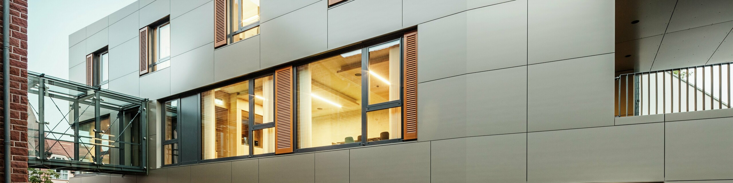 Moderna fasada gradske vijećnice u Alzenauu u Njemačkoj s PREFA aluminijskim kompozitnim pločama u brončanoj boji, u kombinaciji s griljama i ozelenjenim površinama za sadnju. Kvalitetna aluminijska obloga osigurava elegantan i dugotrajan izgled te je idealna za održive građevinske projekte. Ovdje je bočna strana jasno vidljiva.