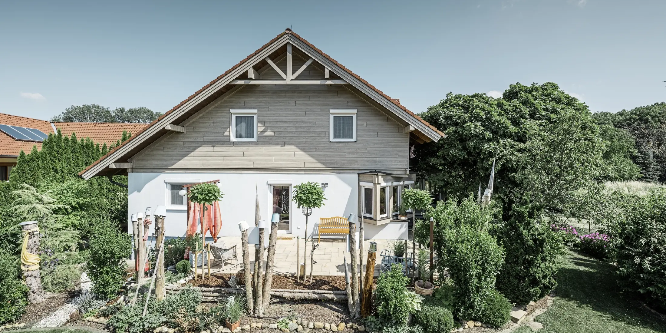 Enkeltfamiliehus med renoveret facade og ny gavlbeklædning i et moderne trælook i grå-beige, kombineret med pudset facade, sadeltag og velplejet have.
