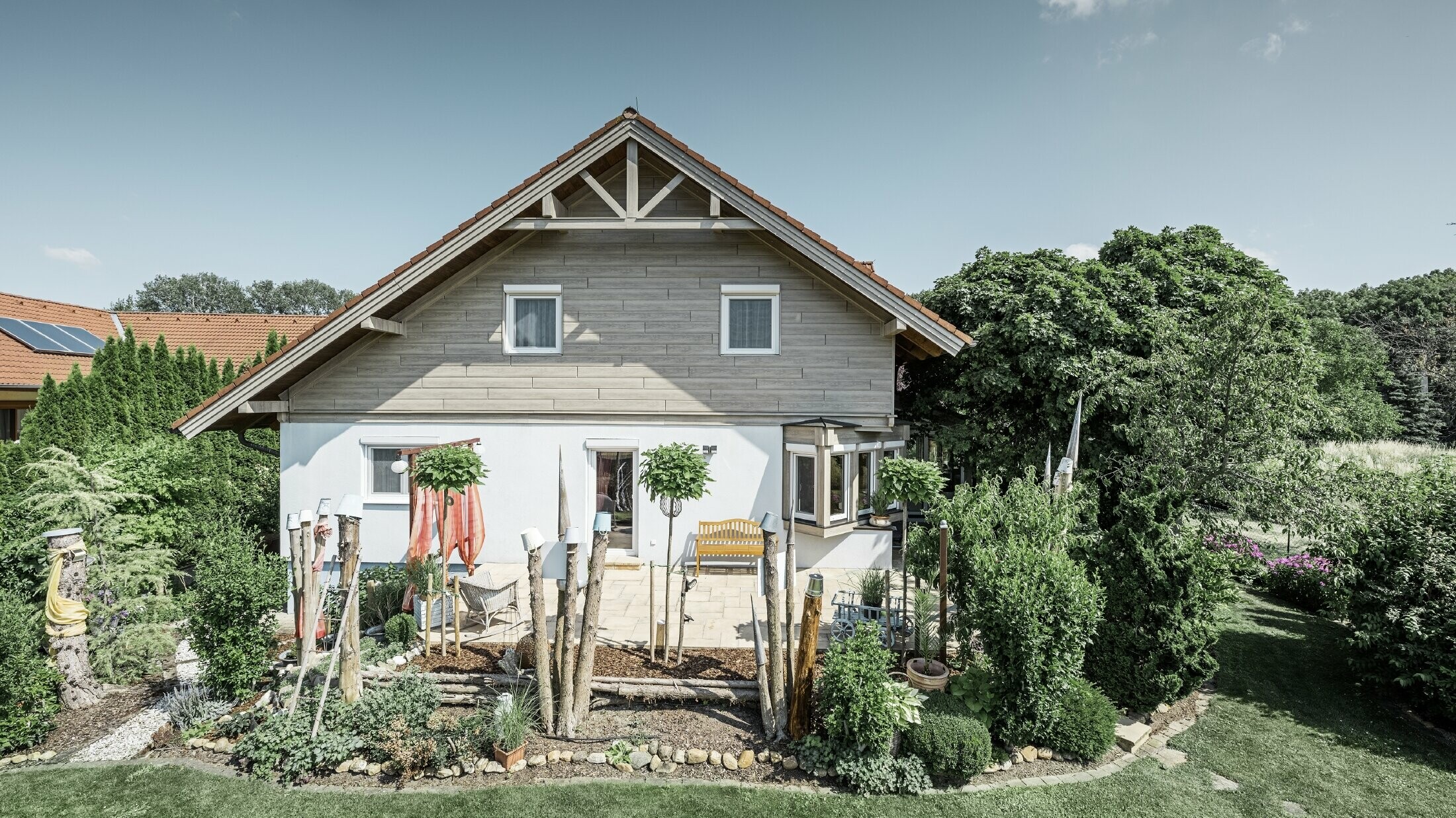 Enkeltfamiliehus med renoveret facade og ny gavlbeklædning i et moderne trælook i grå-beige, kombineret med pudset facade, sadeltag og velplejet have.