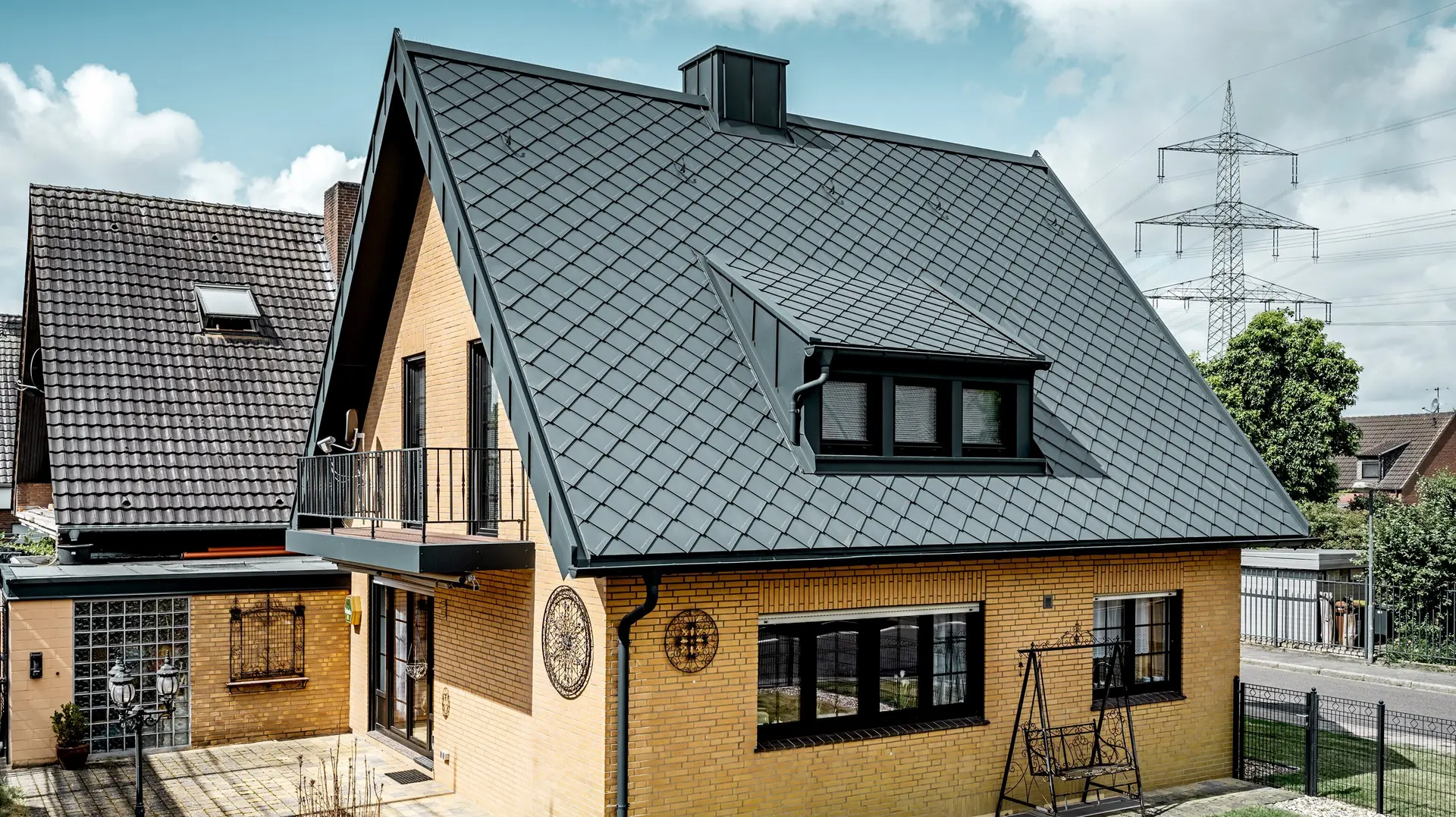Enkeltfamiliehus af gul klinker efter tagrenovering med PREFA tagrombe 29 × 29 i mørk grå; nyt tag med stejlt sadeltag med rammemønster, integreret tagkvist og skorsten, terrasse og have i forgrunden.